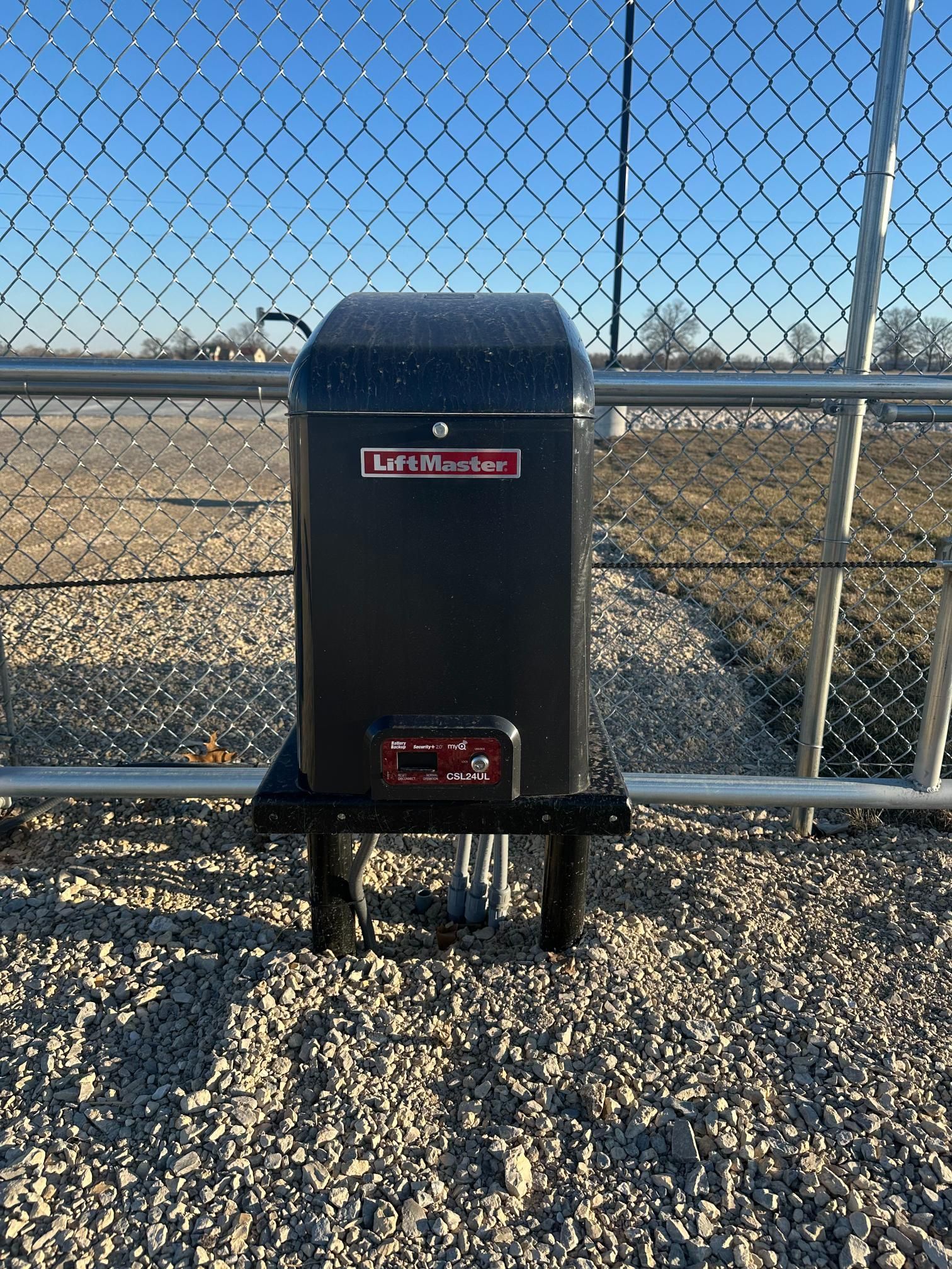 Black gate opener box on a gravel surface next to a chain link fence. 