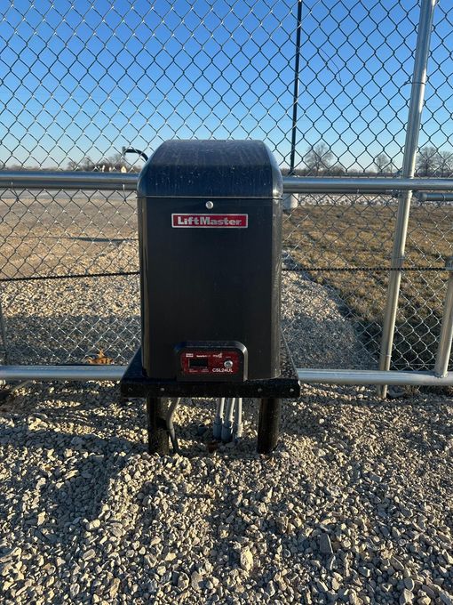 Black gate opener box on a gravel surface next to a chain link fence. 