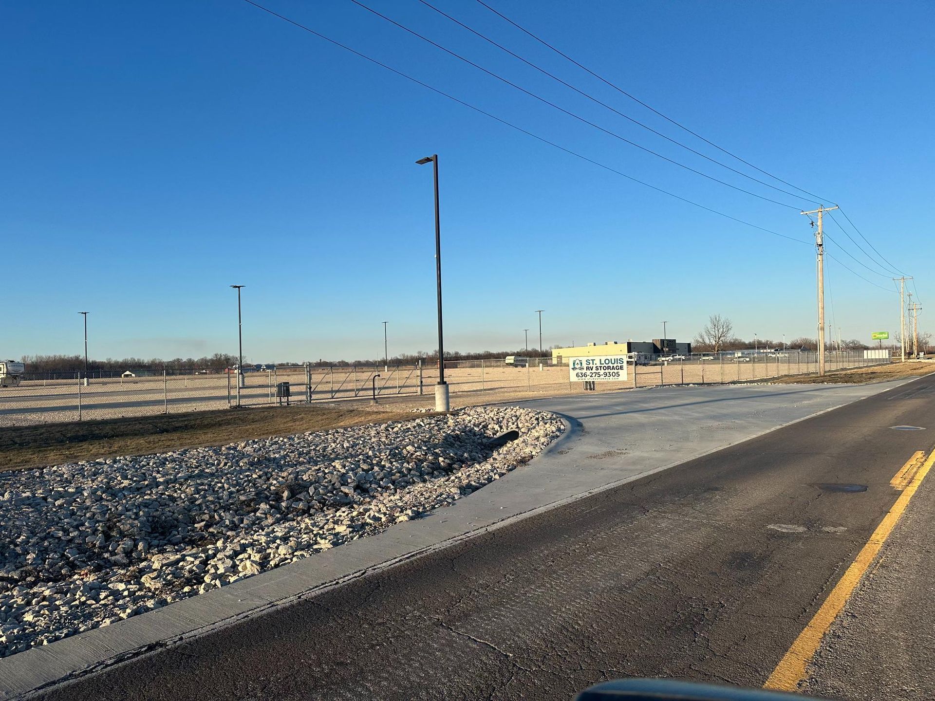 Paved area next to a rocky slope and road, leading towards a vacant lot with a building in the distance.