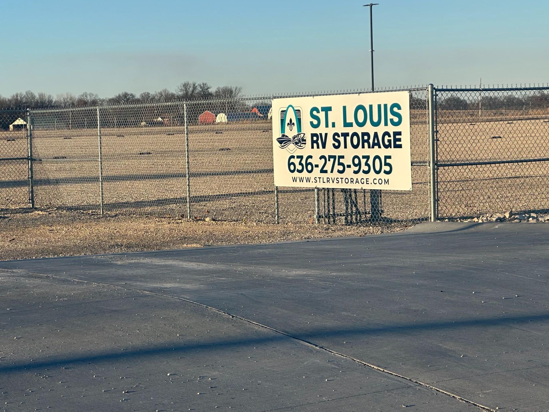 A white sign for St. Louis RV Storage with a phone number and website attached to a chain-link fence by a gravel lot.