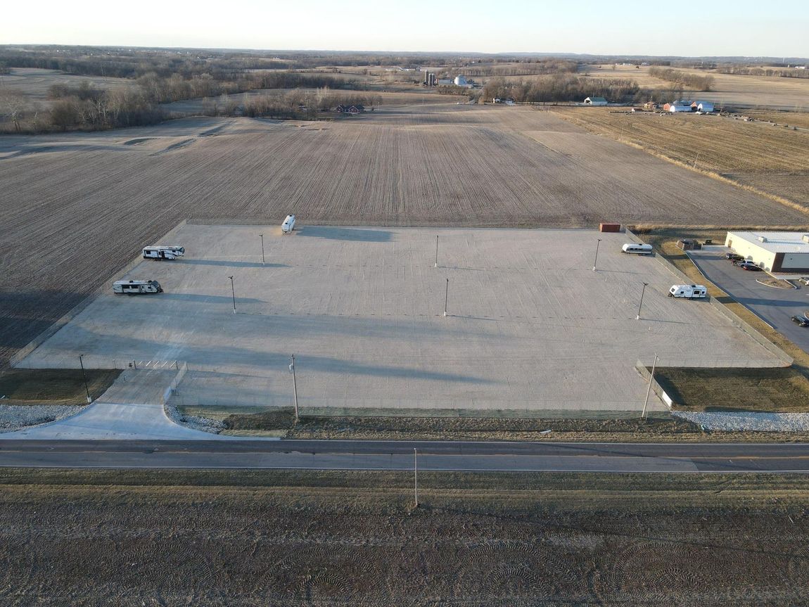 Aerial view of a gravel parking lot with vehicles, next to a building and a harvested field.