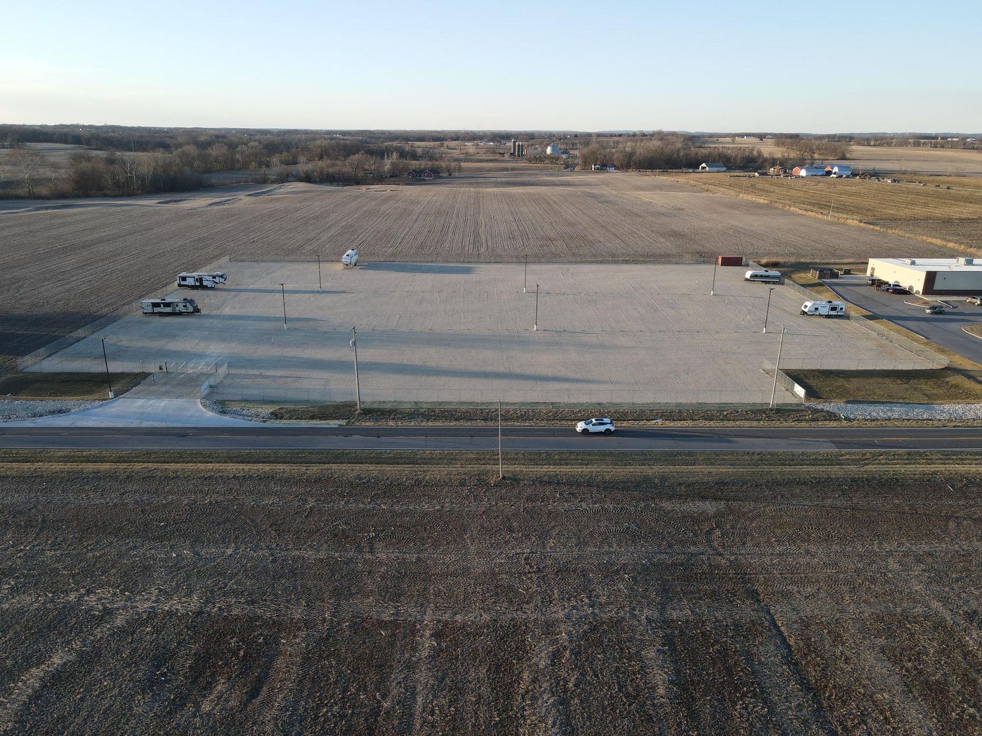 An aerial view of a rectangular gravel lot surrounded by harvested fields, with a road and a building nearby.