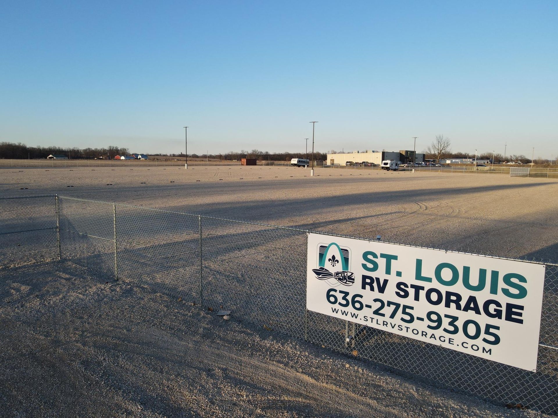 A gravel RV storage lot with a St. Louis RV Storage sign on a chain-link fence under a clear, bright blue sky.