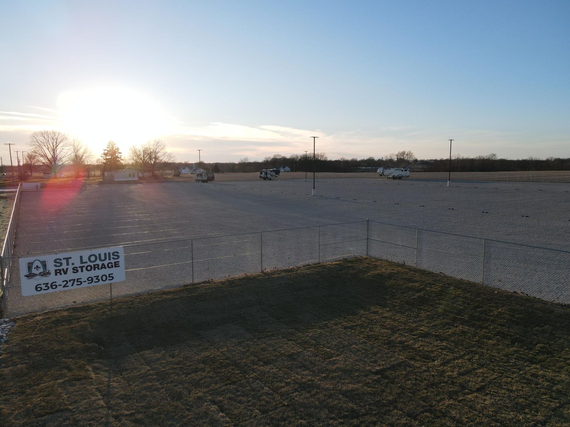Gravel lot with chain-link fence, sign, and distant vehicles under a bright sun.