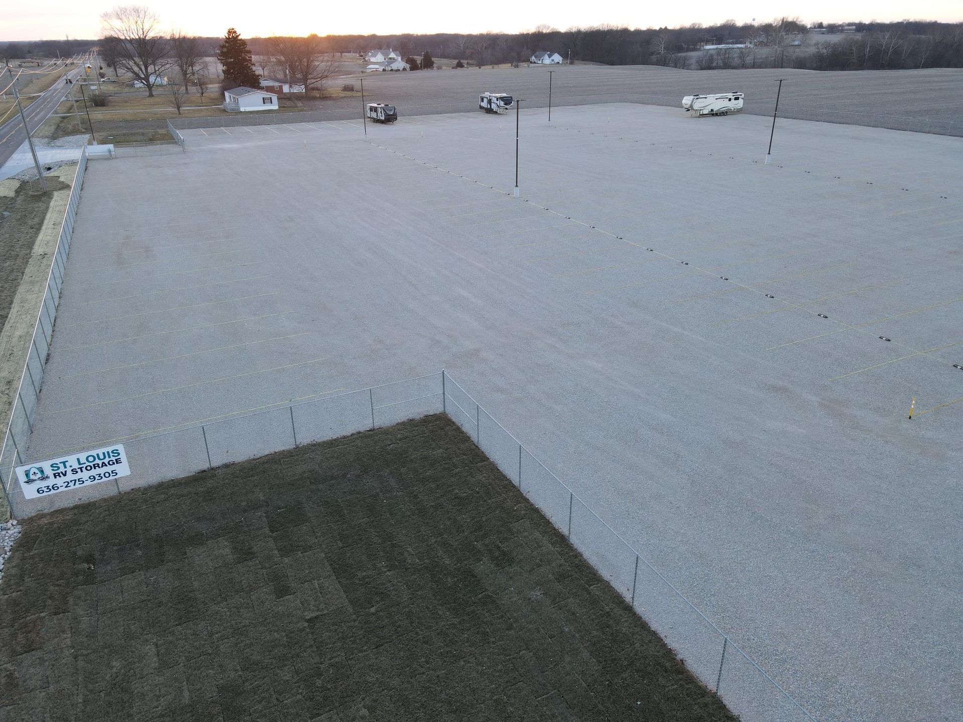 Gravel lot with vehicles, grass, fence, and a sign. The sky is a gradient of colors.