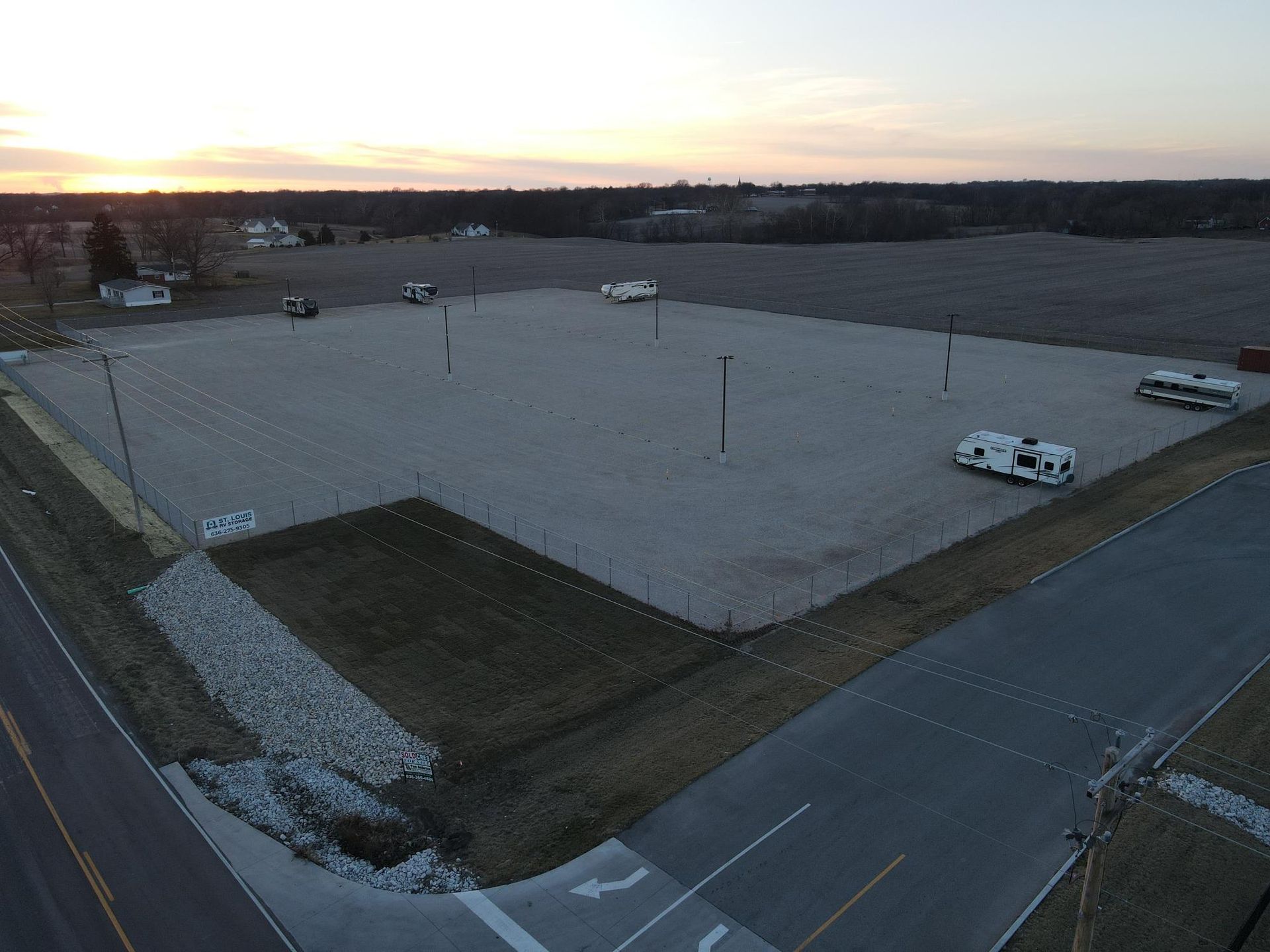 Gravel lot with RVs, light poles, and a grassy area. Road in foreground, fields in background under a sunset.