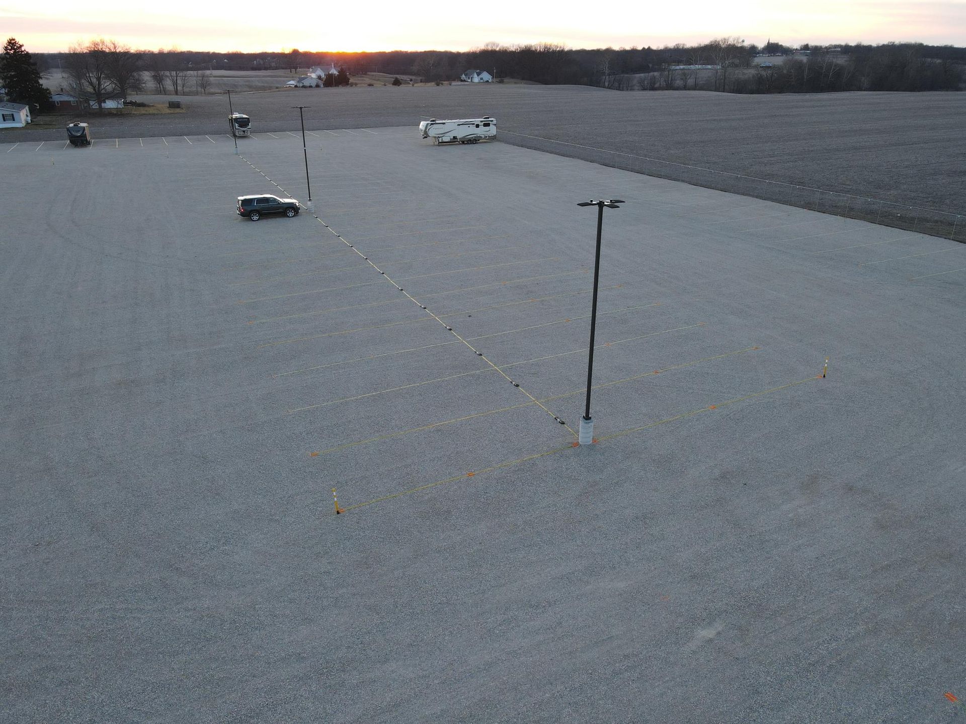 Gravel lot with a black vehicle and light poles; field and buildings in background.