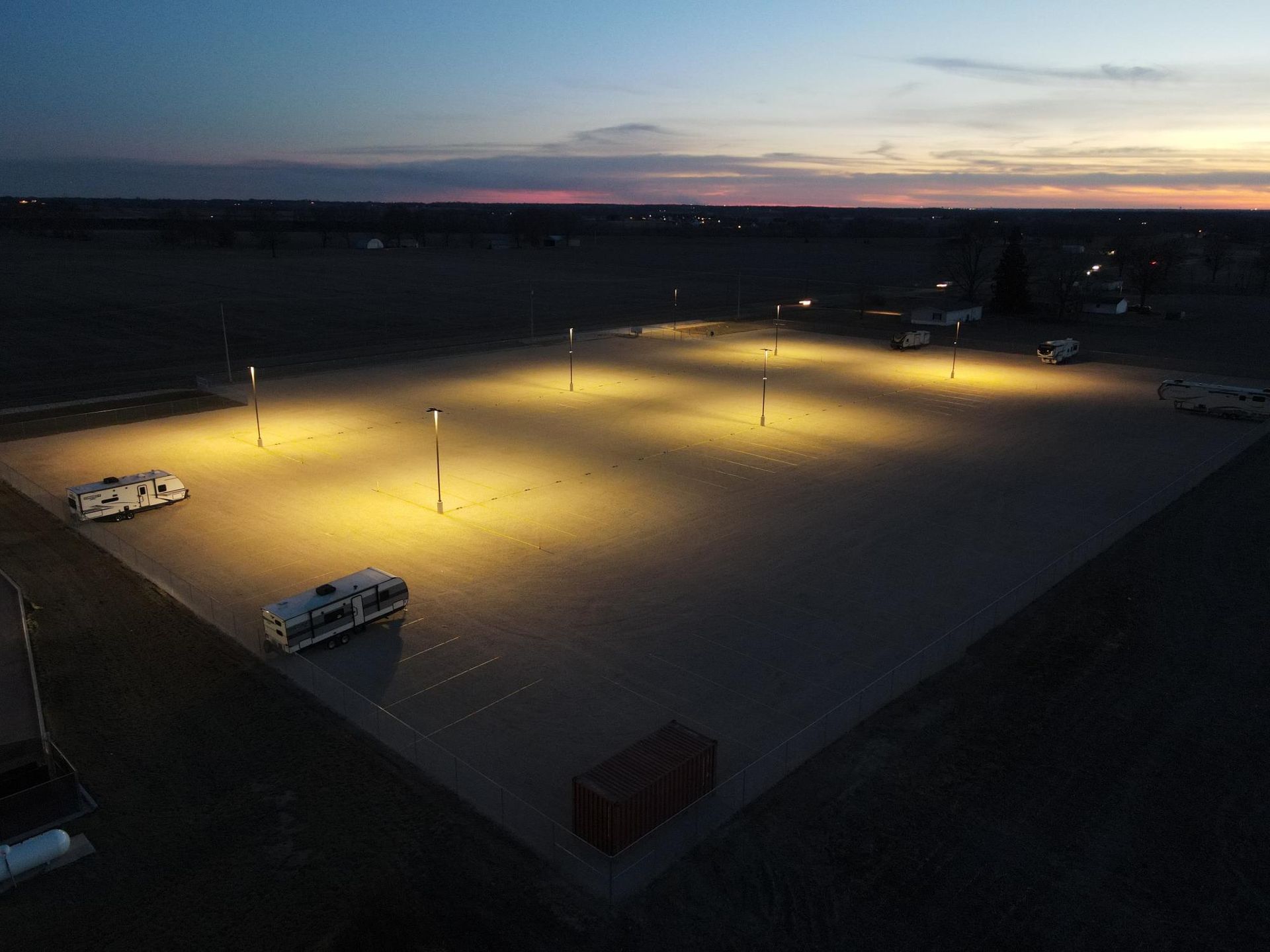 Aerial view of an empty gravel parking lot at dusk, lit by several tall streetlights with a few parked RVs.
