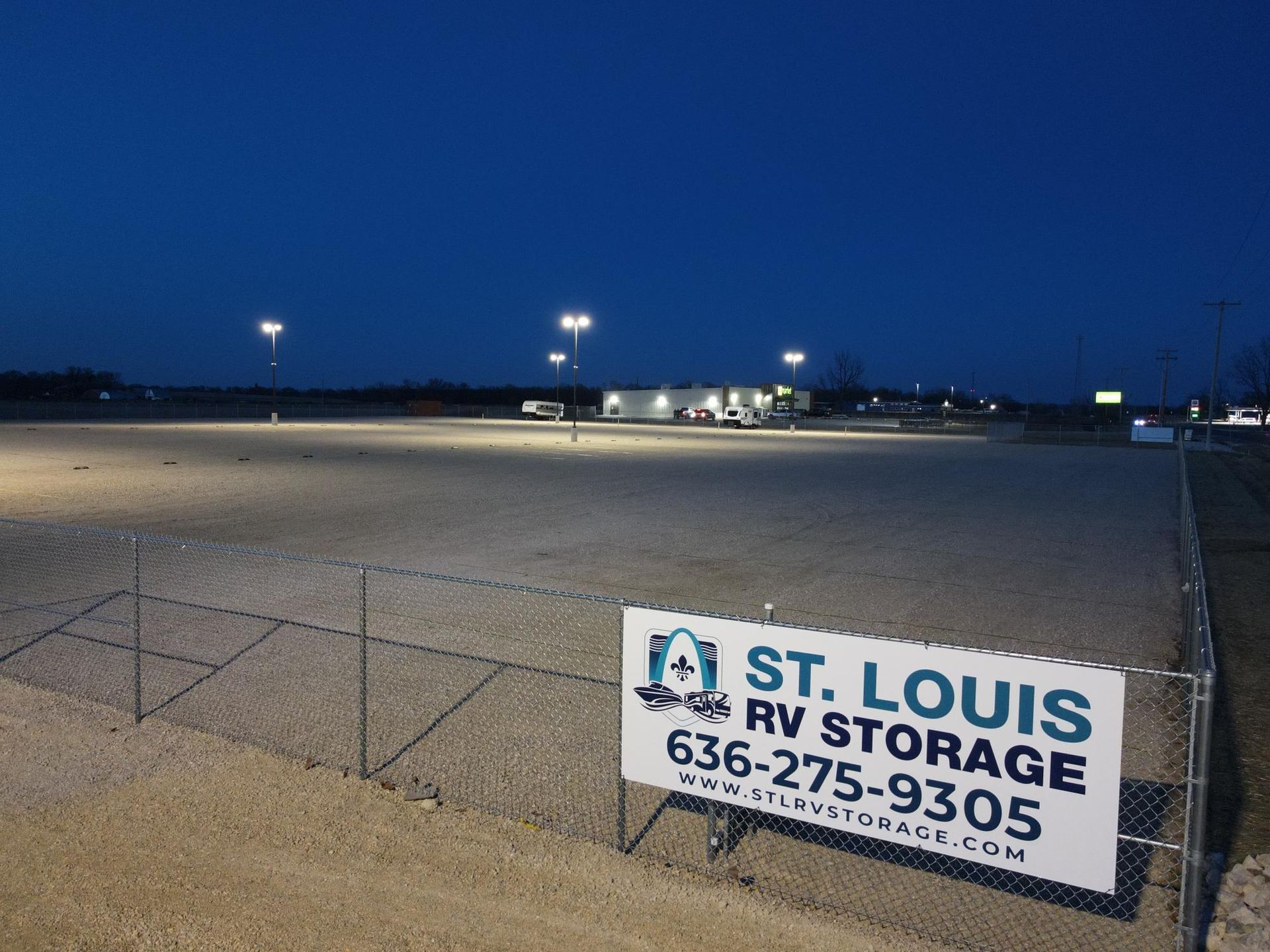 Gravel parking lot at dusk with sign for St. Louis RV Storage; lights illuminate the area.