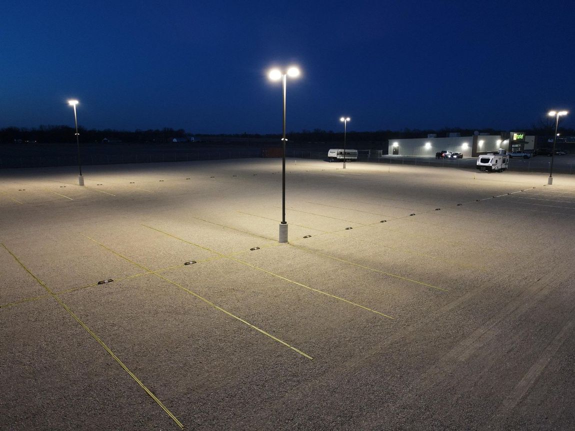 Nighttime view of a vacant, gravel parking lot illuminated by tall street lights. A few vehicles are parked in the distance.