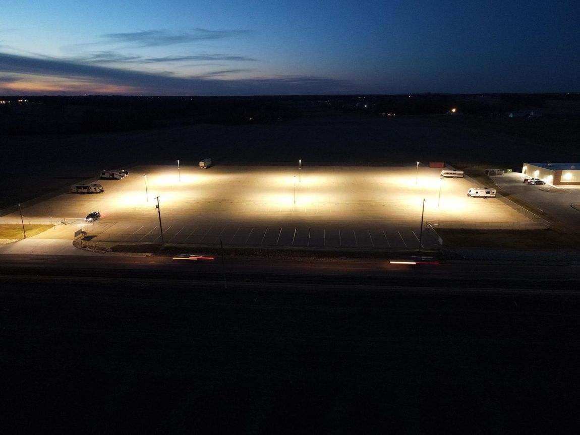 Parking lot lit by tall streetlights at dusk. Cars parked. Dark sky in the background.