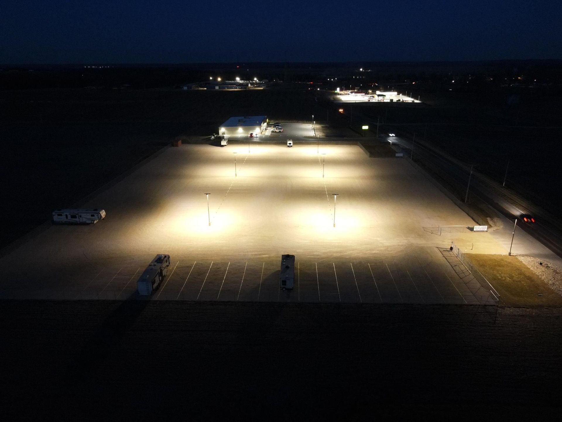 Night aerial view of a brightly lit parking lot with a building and parked vehicles.