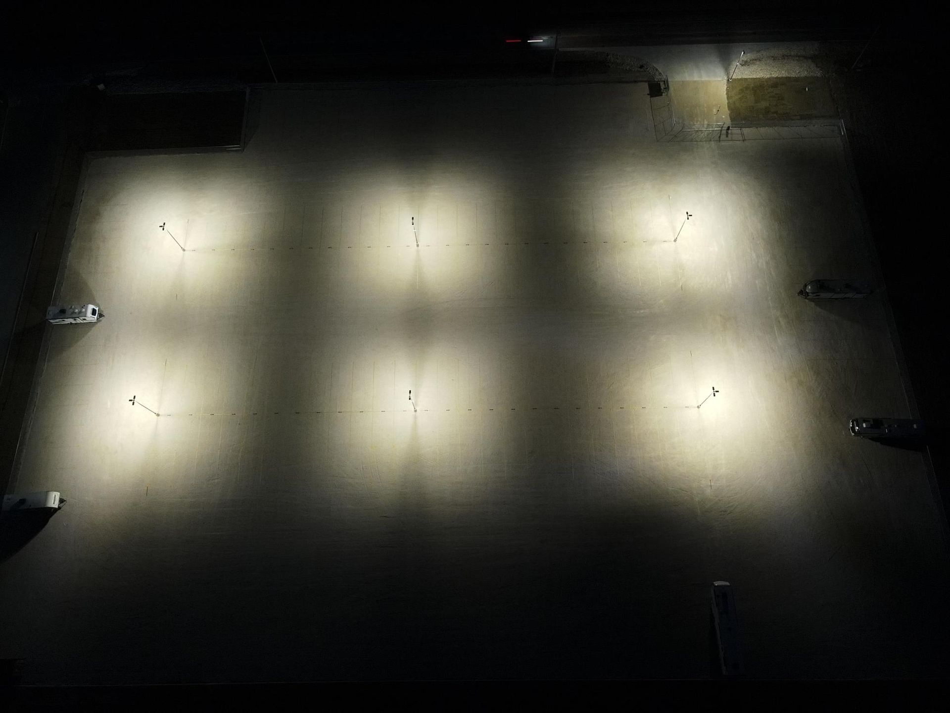 Overhead view of a dimly lit parking lot at night with six bright spotlights illuminating the pavement and scattered vehicles.