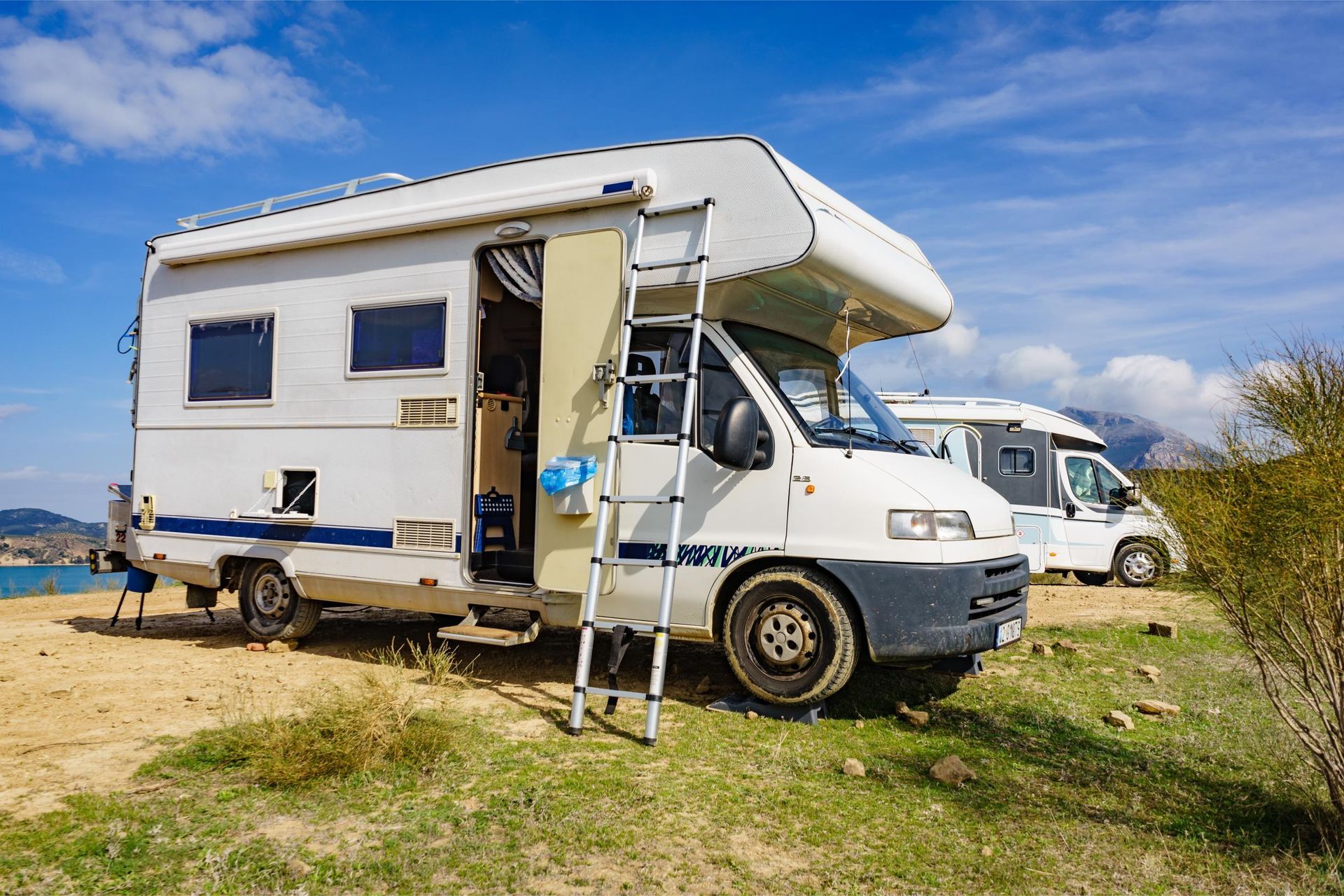 White RV parked on a grassy area, blue sky. Ladder to open door. Another RV in the background.