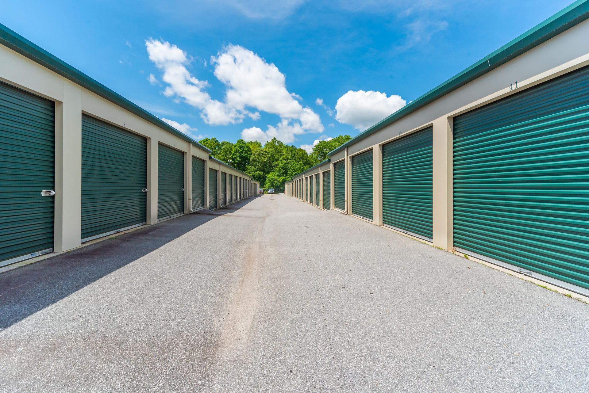 Rows of green storage unit doors line a gravel path under a blue sky with clouds.