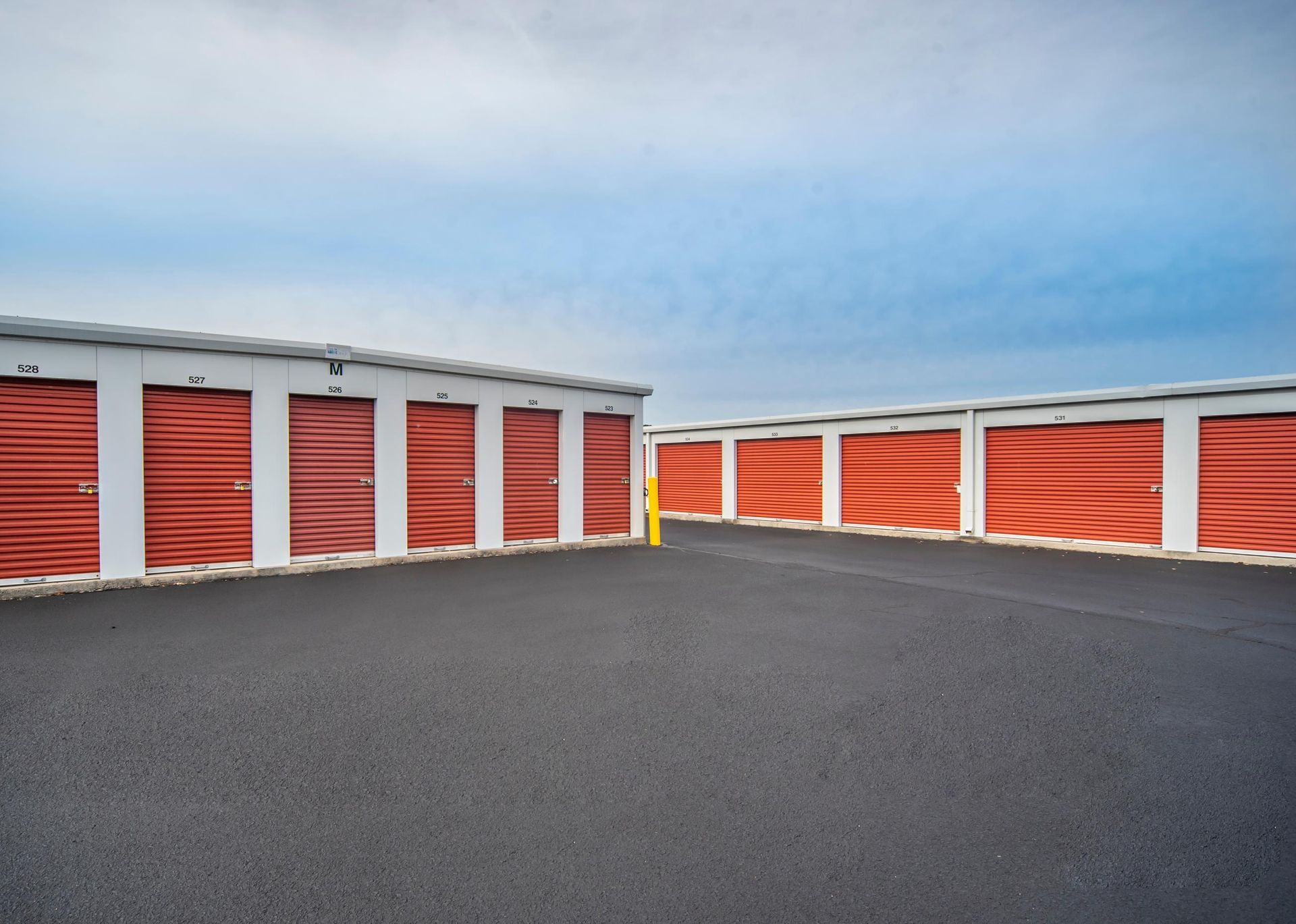 Storage units with red doors and white trim, arranged on a black asphalt lot, under a blue sky.