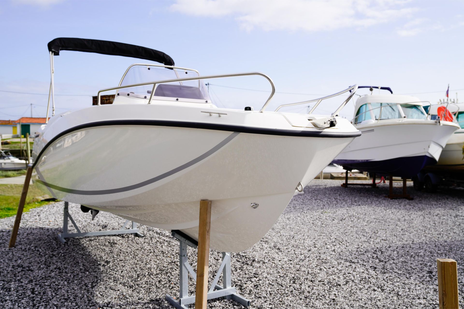 White boat on supports, parked on gravel. Another boat in background, blue hull. Sunny day.