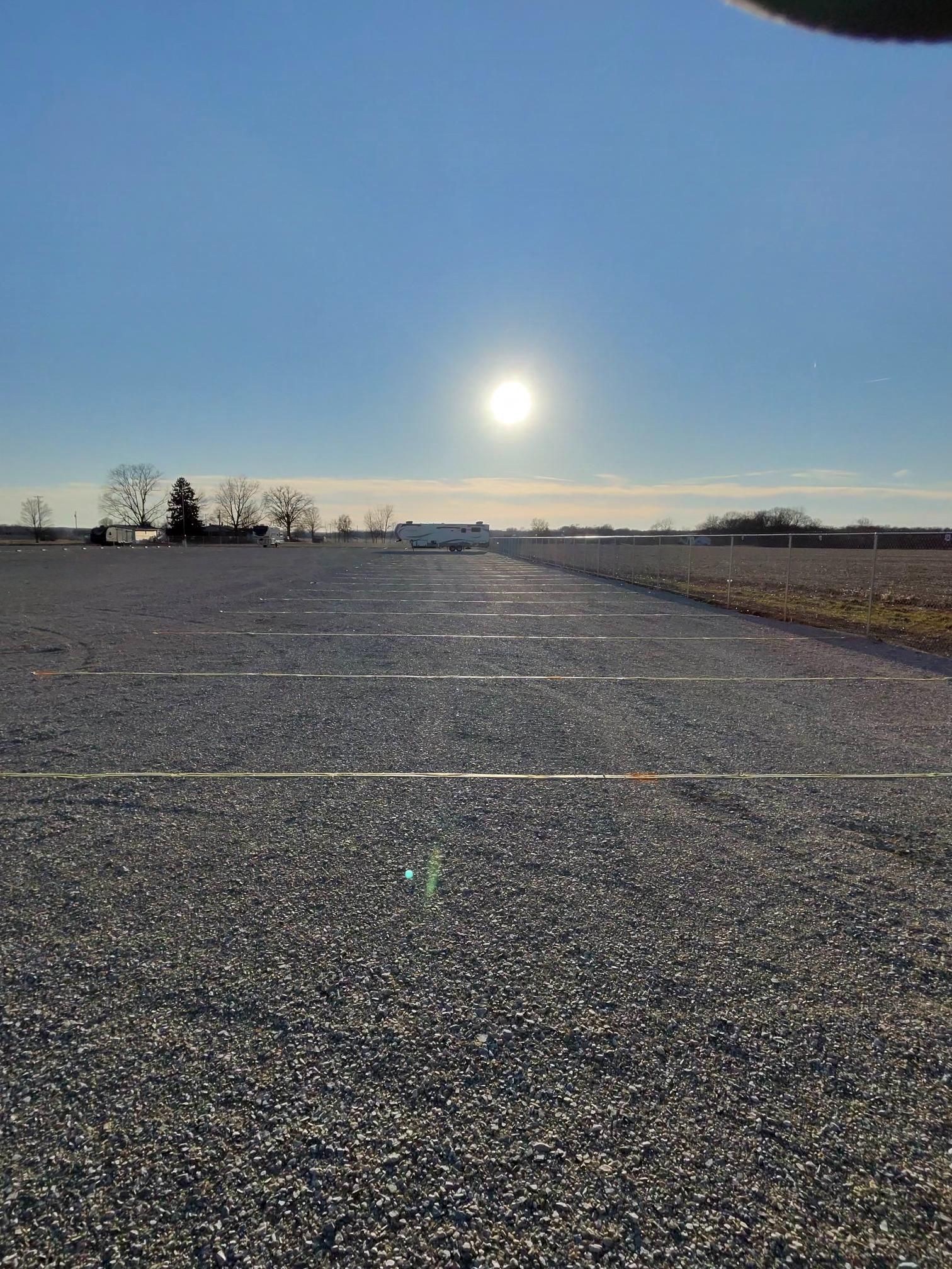 Gravel parking lot under a bright sun. Distant buildings and trees on the horizon.