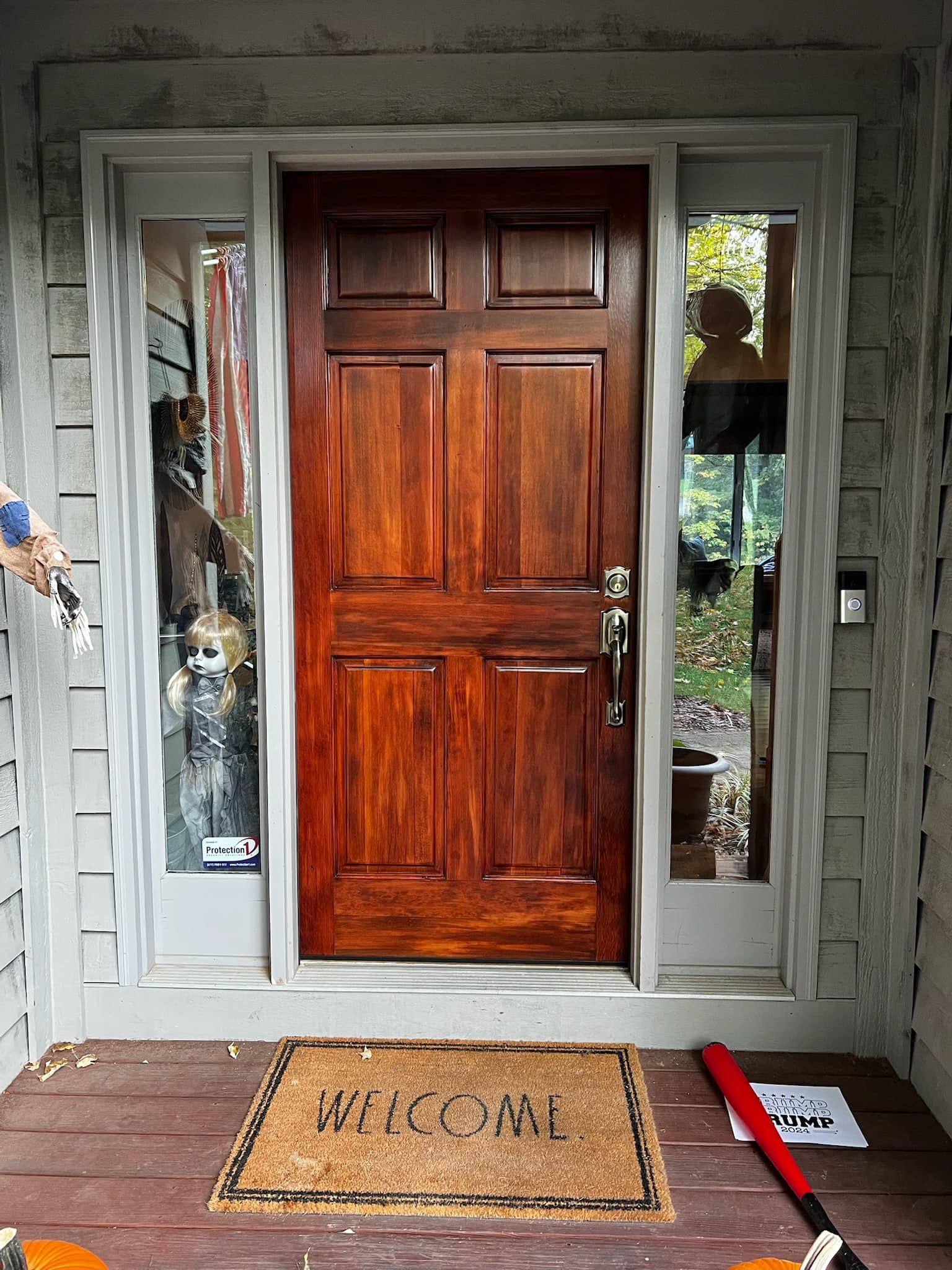 Wooden front door with sidelights. Welcome mat on porch, flanked by gray siding.