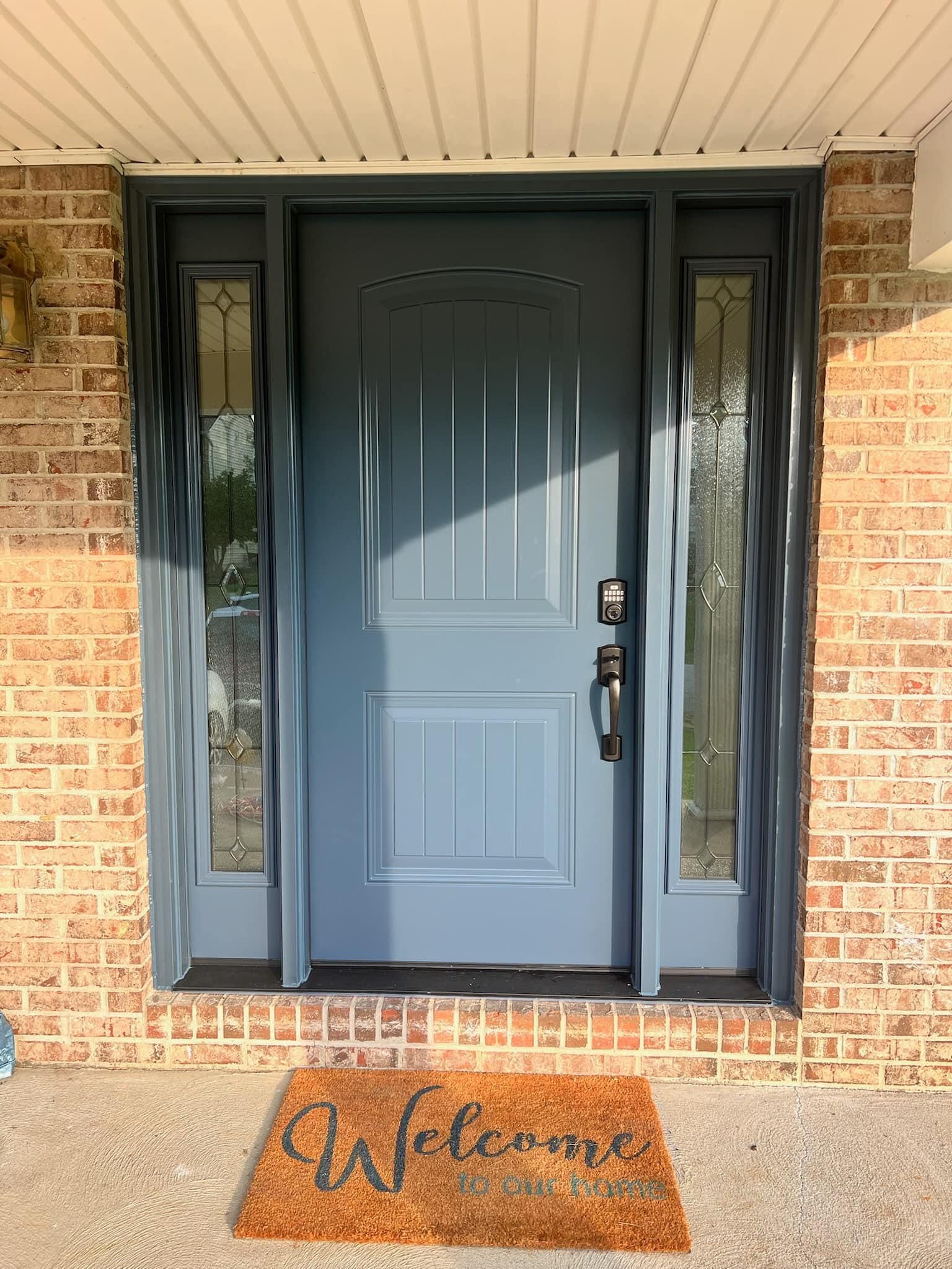 Blue front door with sidelights, welcome mat on the porch, brick exterior.