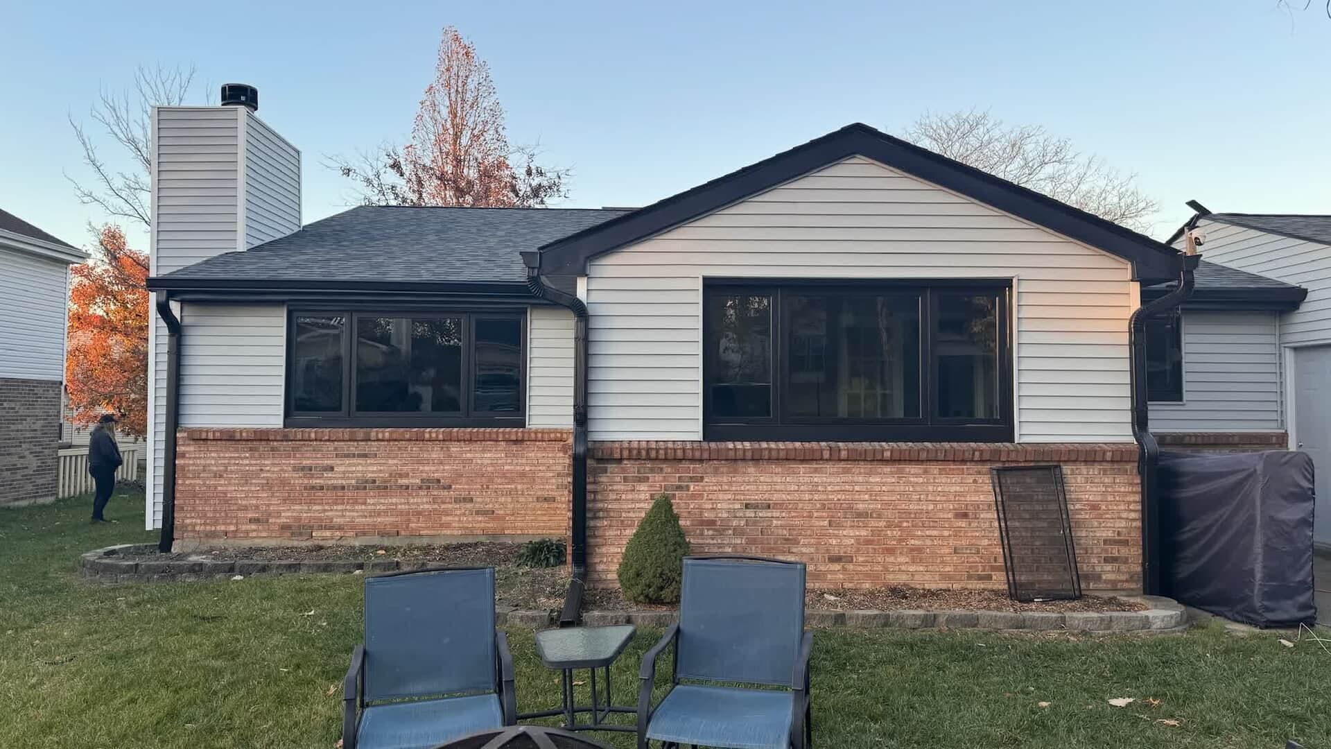 Backyard of a home with brick base, gray siding, black-framed windows, and two chairs on the lawn.