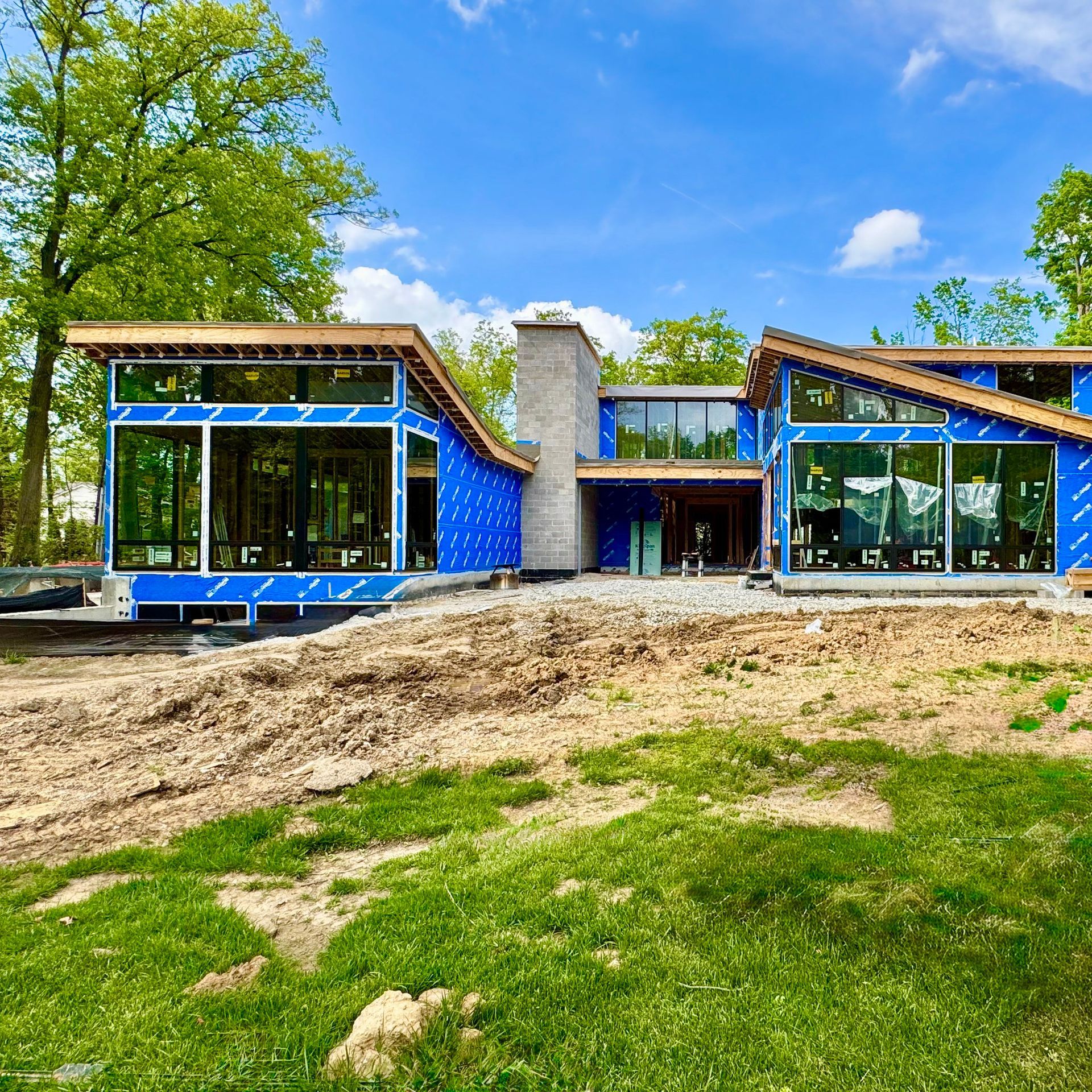 Modern house under construction with large windows, blue wrap, and dirt foreground.