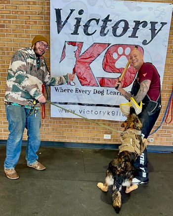 Two men are standing next to a dog in front of a sign that says victory k9