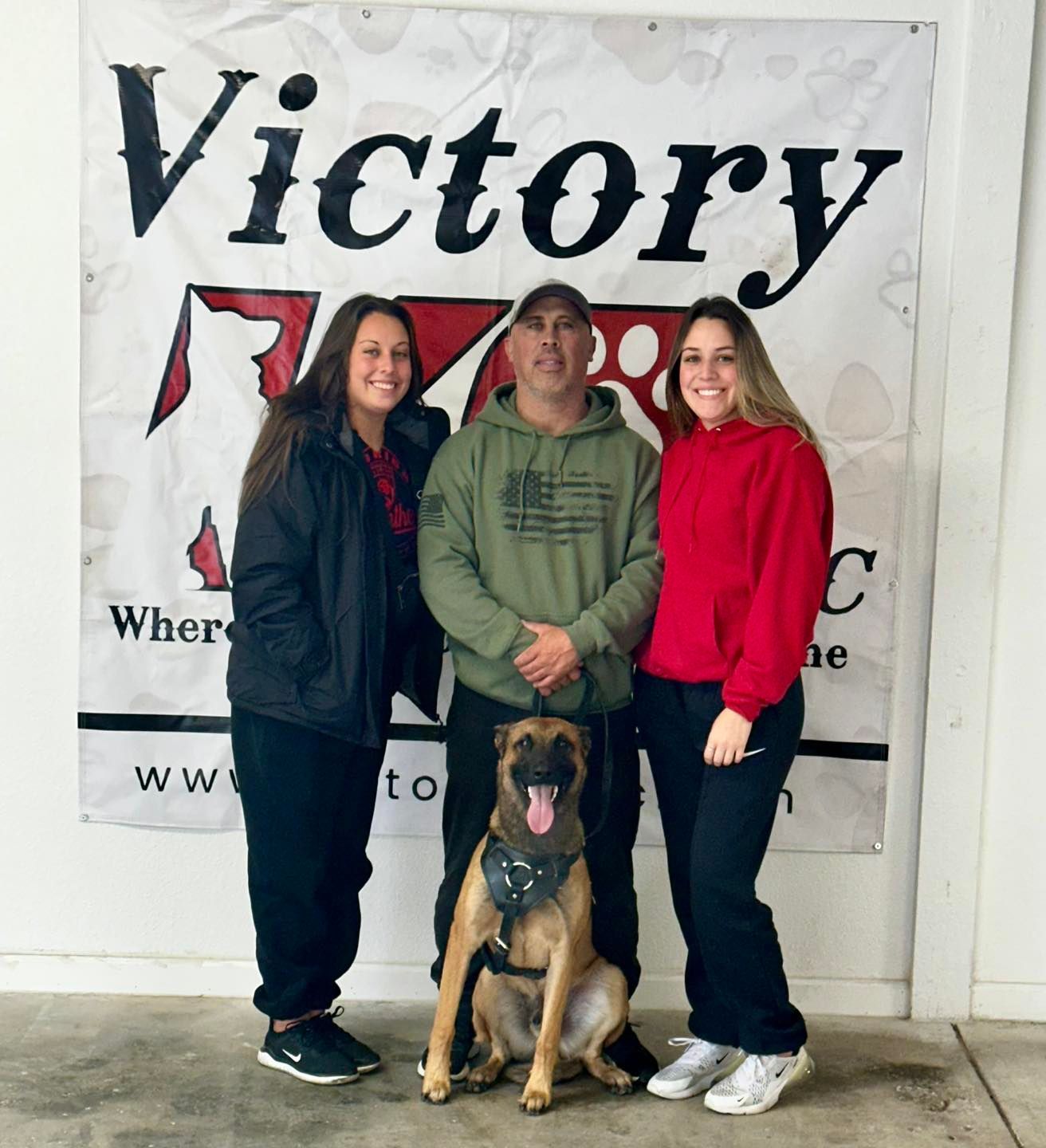 A man and two women pose with a dog in front of a sign that says victory k9