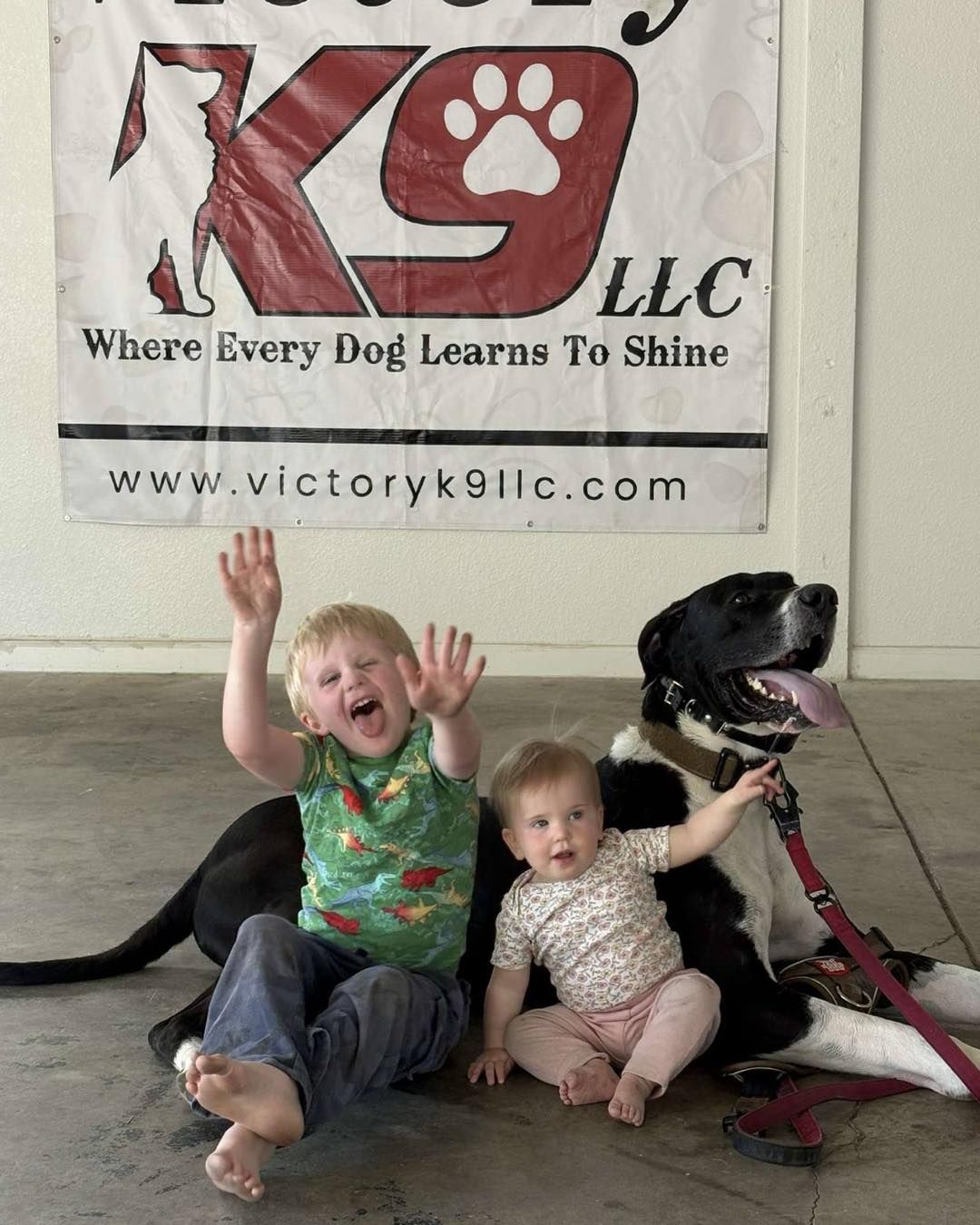 Two children sitting next to a dog in front of a victory k9 banner