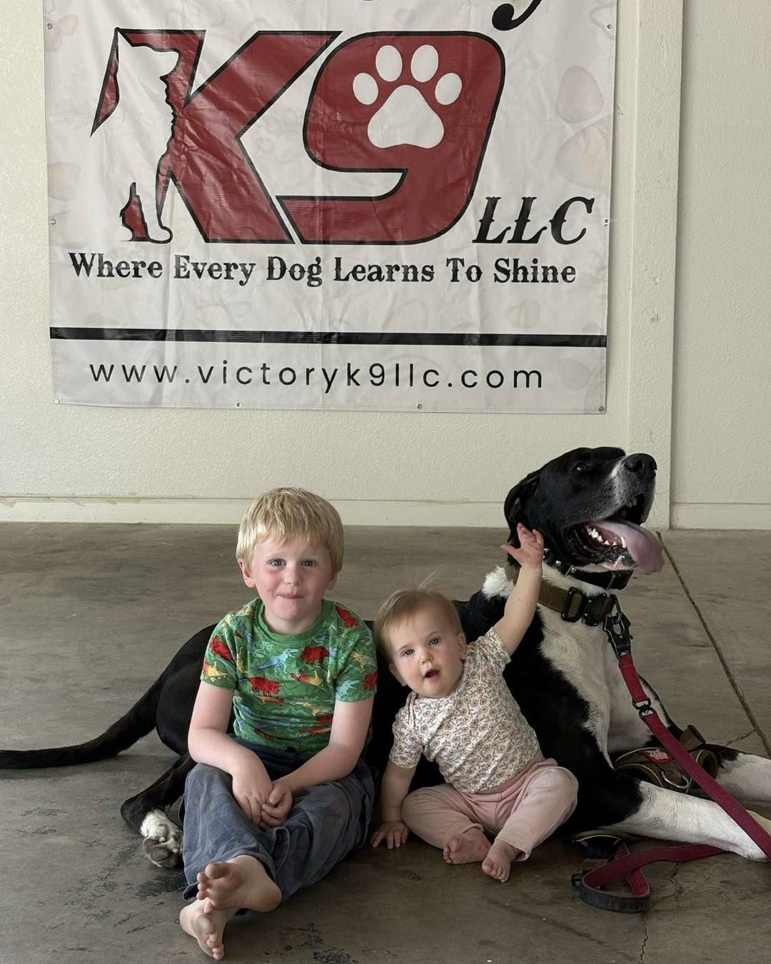 Two children are sitting next to a dog in front of a sign that says k9