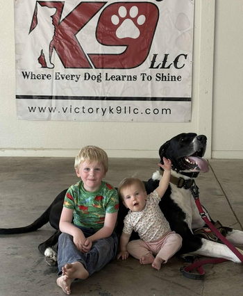 Two children are sitting next to a dog in front of a sign that says k9