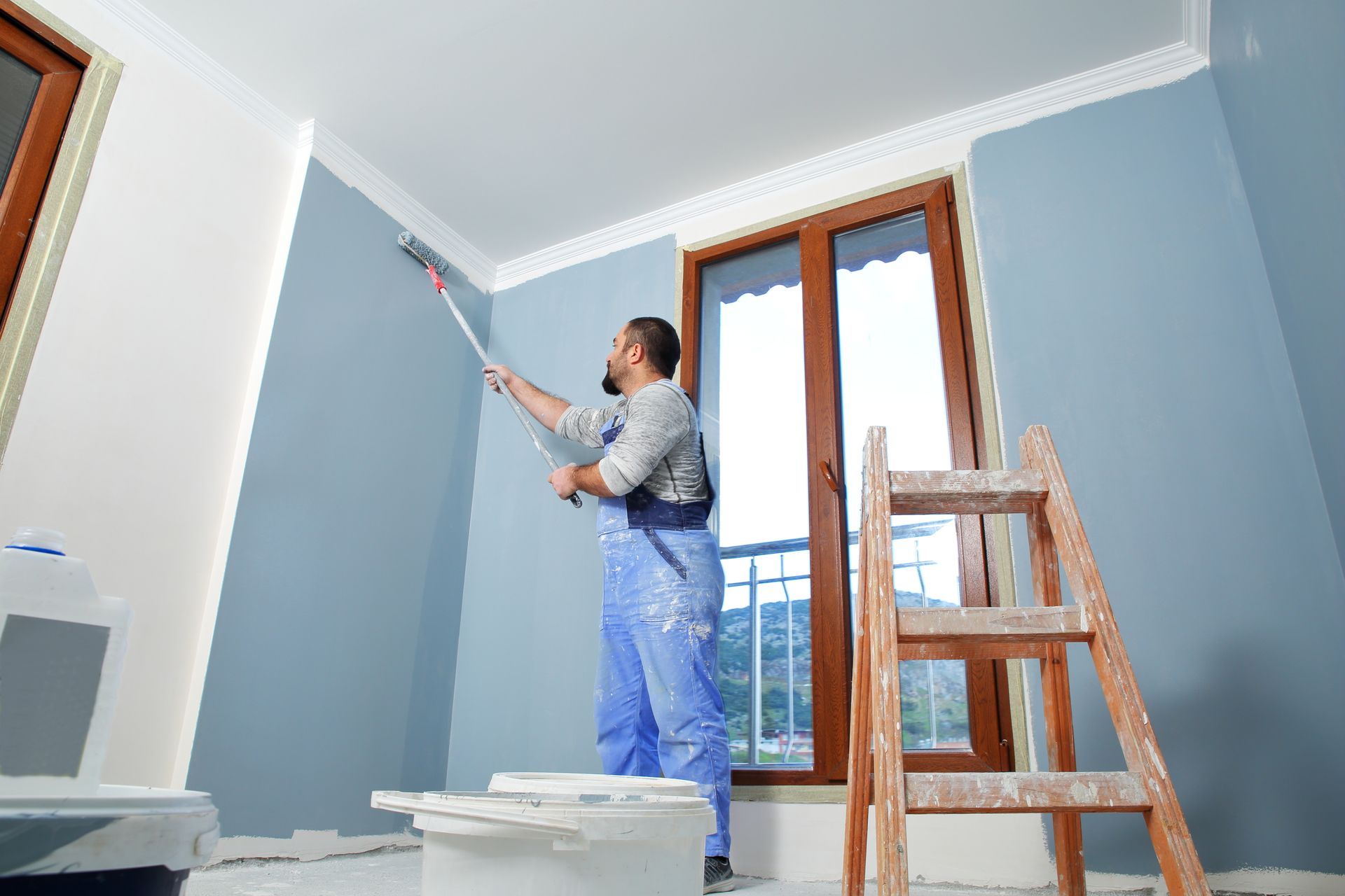 Man in blue overalls painting a wall blue with a roller. Wooden ladder and window.