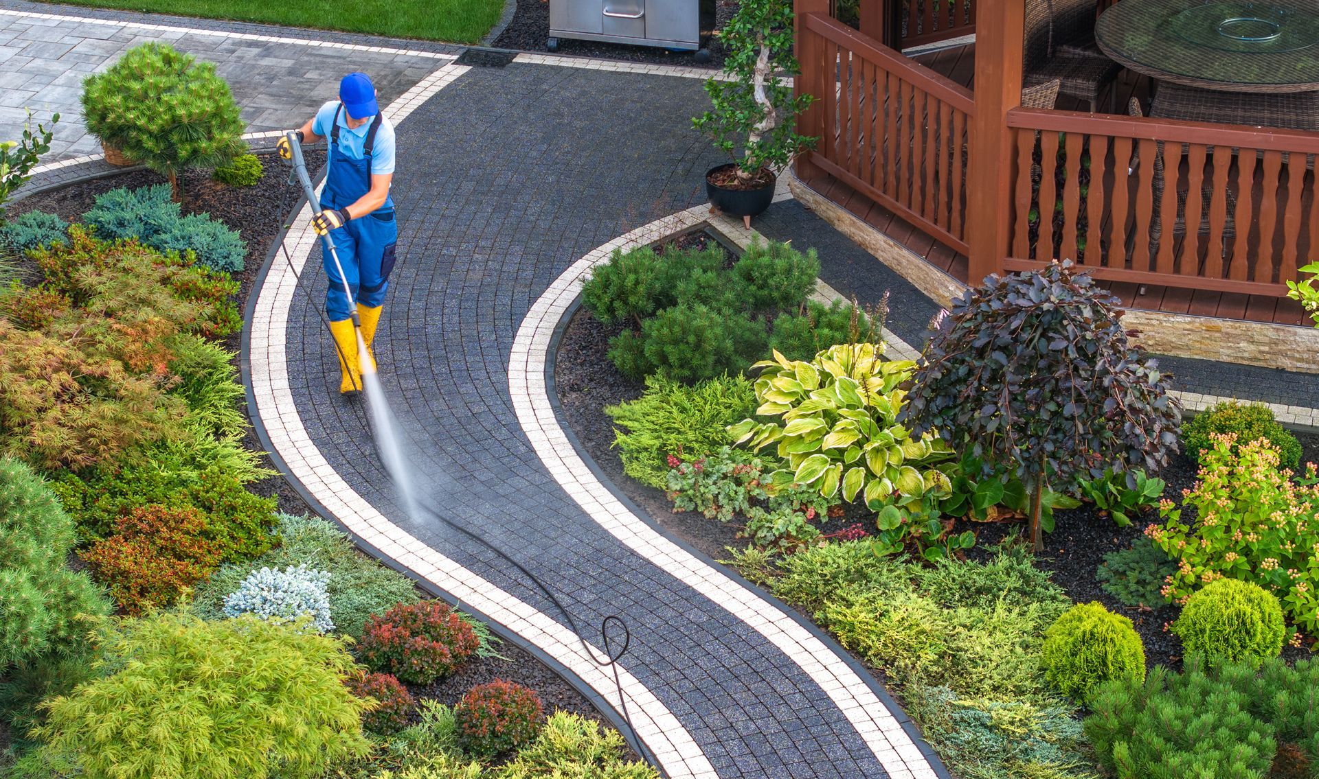 Man in blue suit power washing a winding black stone path edged with white, surrounded by greenery.