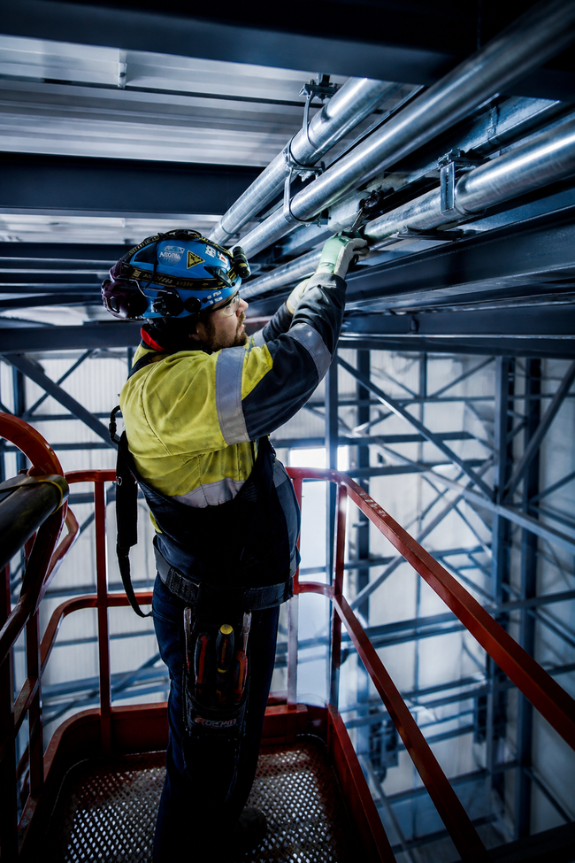 Worker in safety gear inspecting overhead pipes on a metal platform in an industrial facility