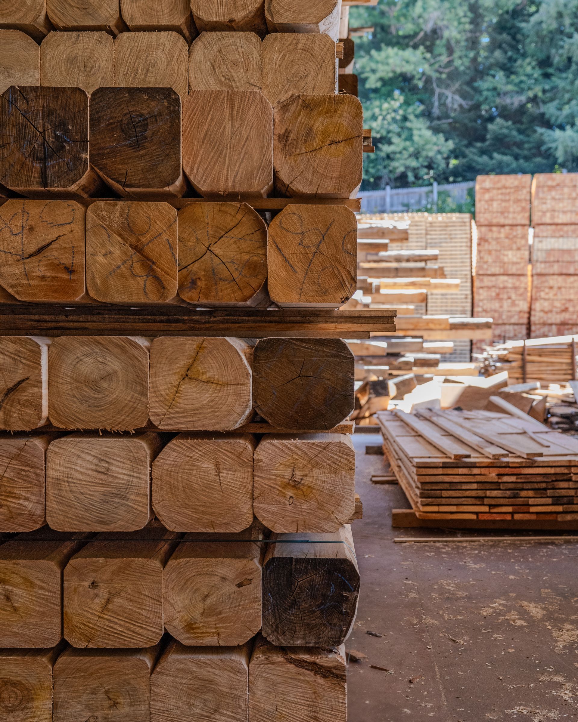 Catasta di legname tagliato a quadrotti in un deposito di legname, con altre cataste di legna visibili sullo sfondo.