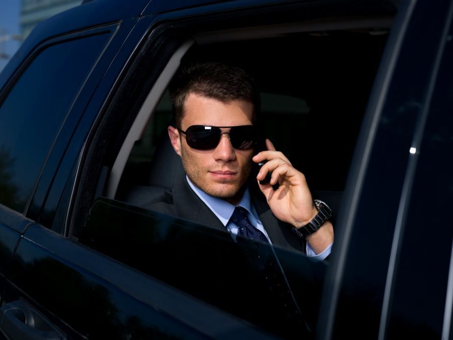 Man in sunglasses, in a car, talking on the phone. Wearing suit, dark interior.