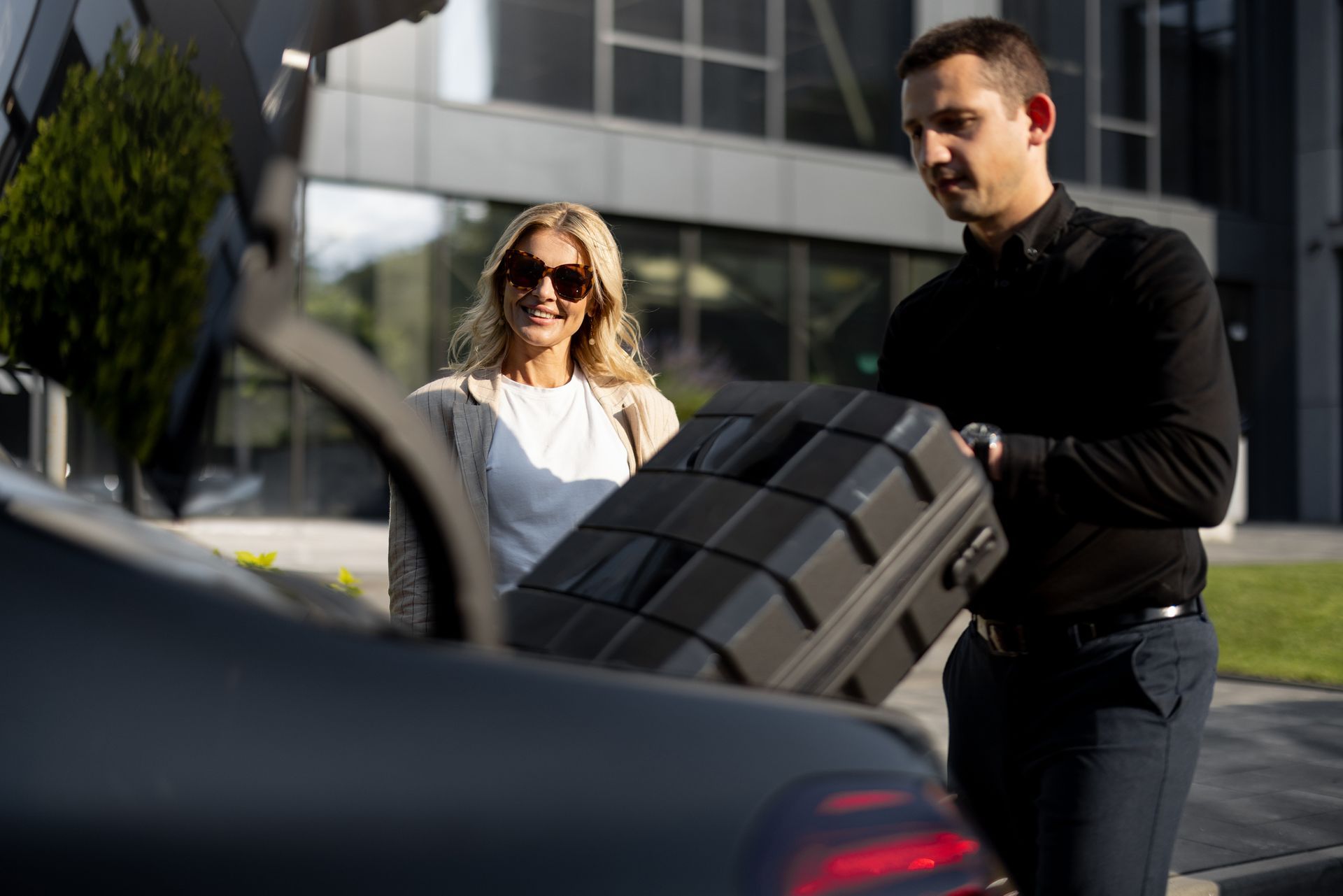 Man loading a black suitcase into a car trunk while a woman watches; outdoors.