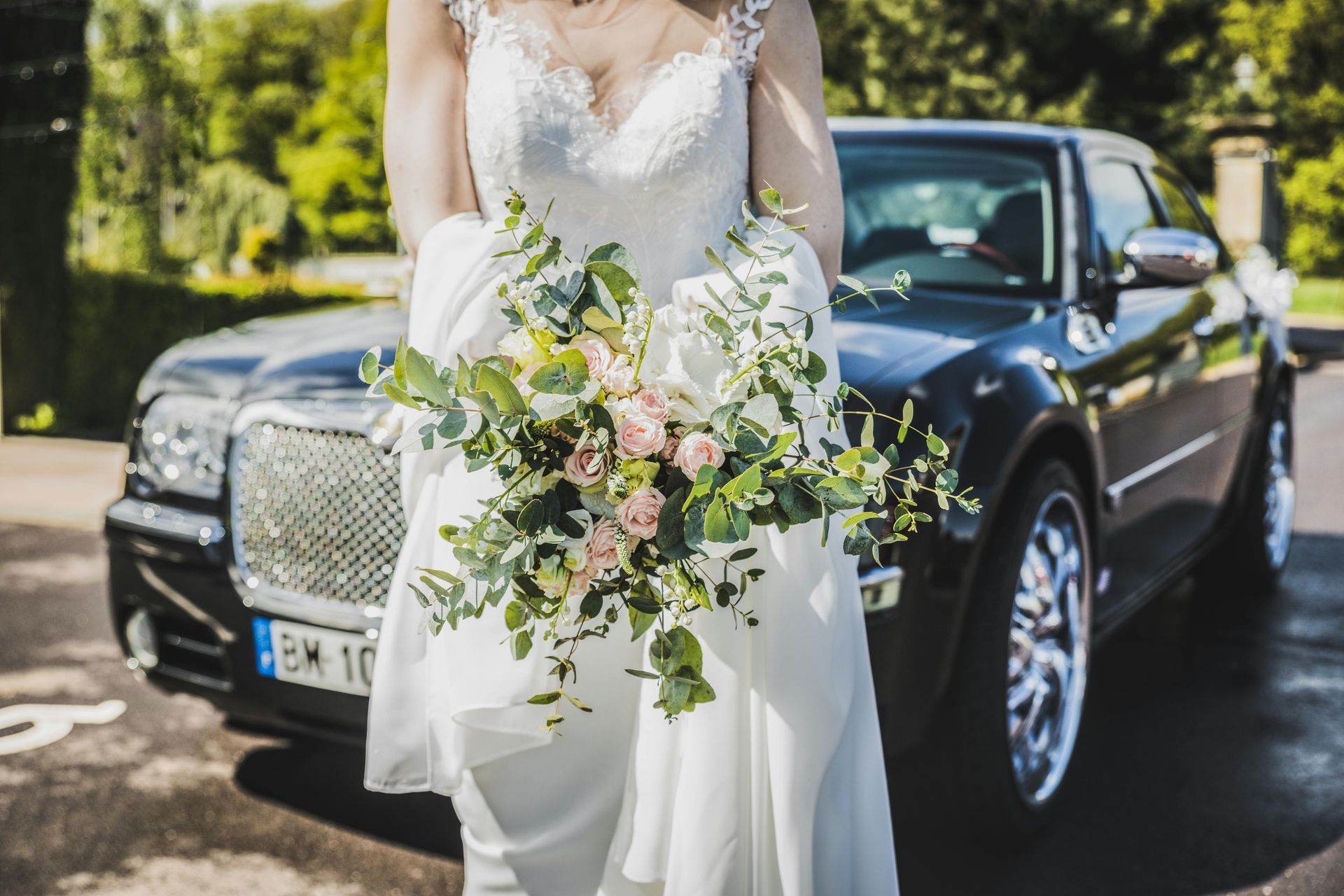 Bride in a white dress holding flowers in front of a black car.