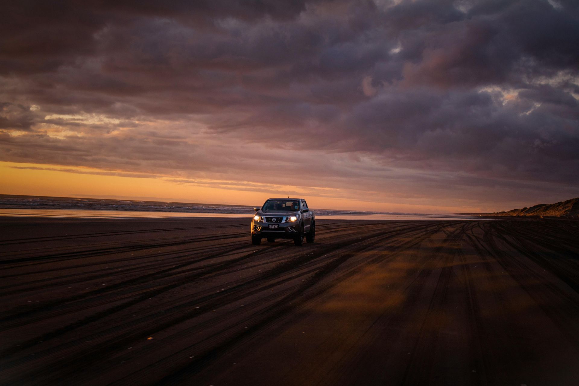 A truck driving on a dark beach at sunset, with orange and purple clouds overhead.