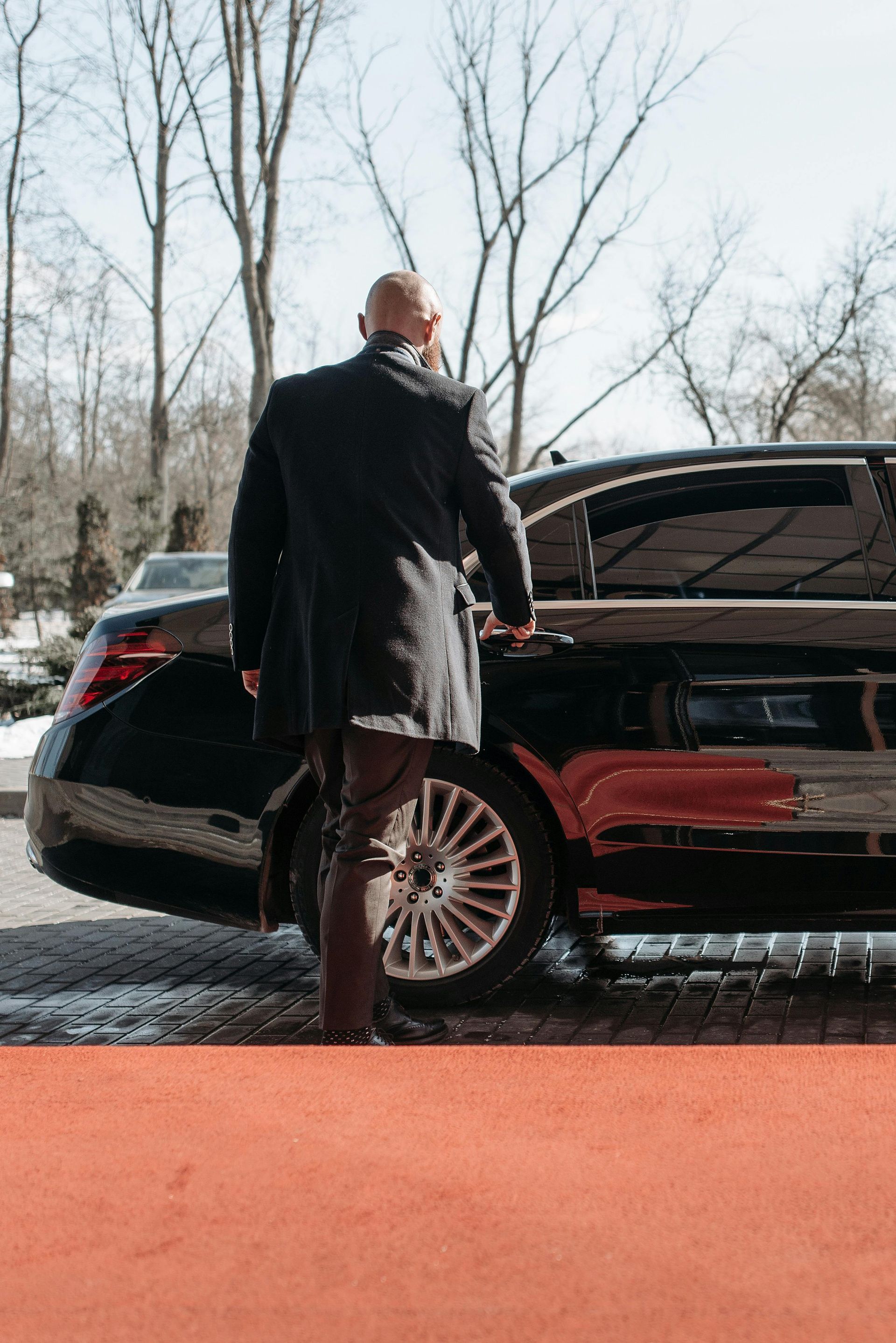 Bald man in suit, getting into a black car on a red carpet.