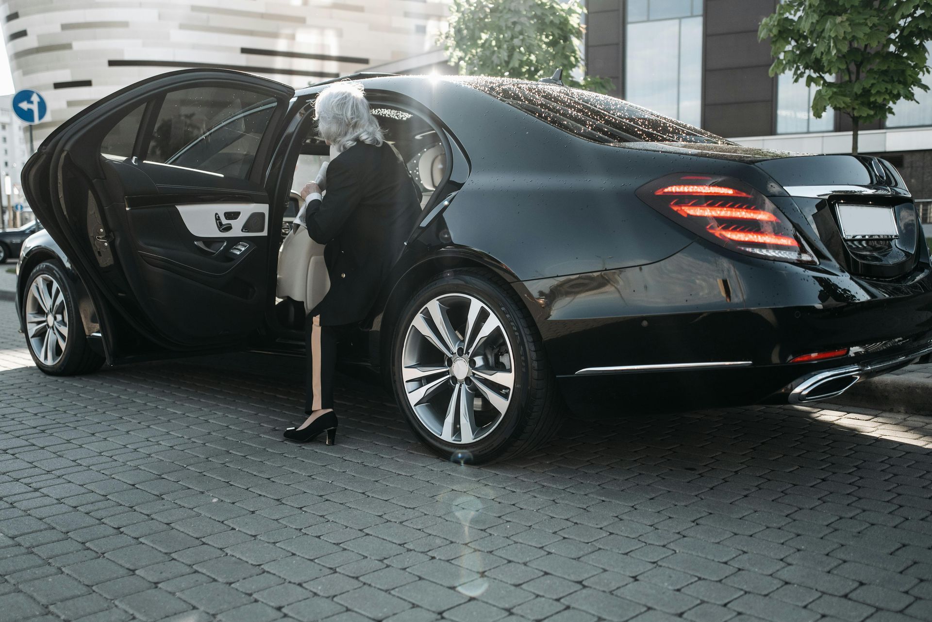 An elderly woman in black attire steps out of a black luxury car on a city street.