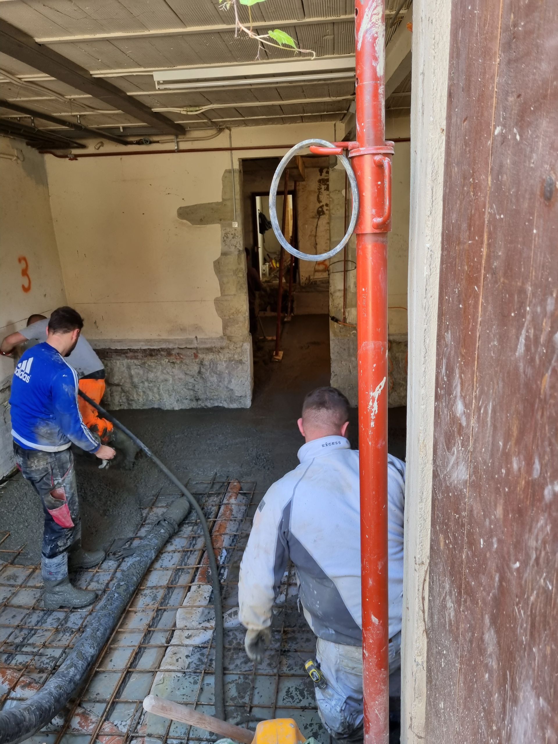 Deux ouvriers coulent et étalent du béton sur une grille métallique à l'intérieur d'une pièce en rénovation.