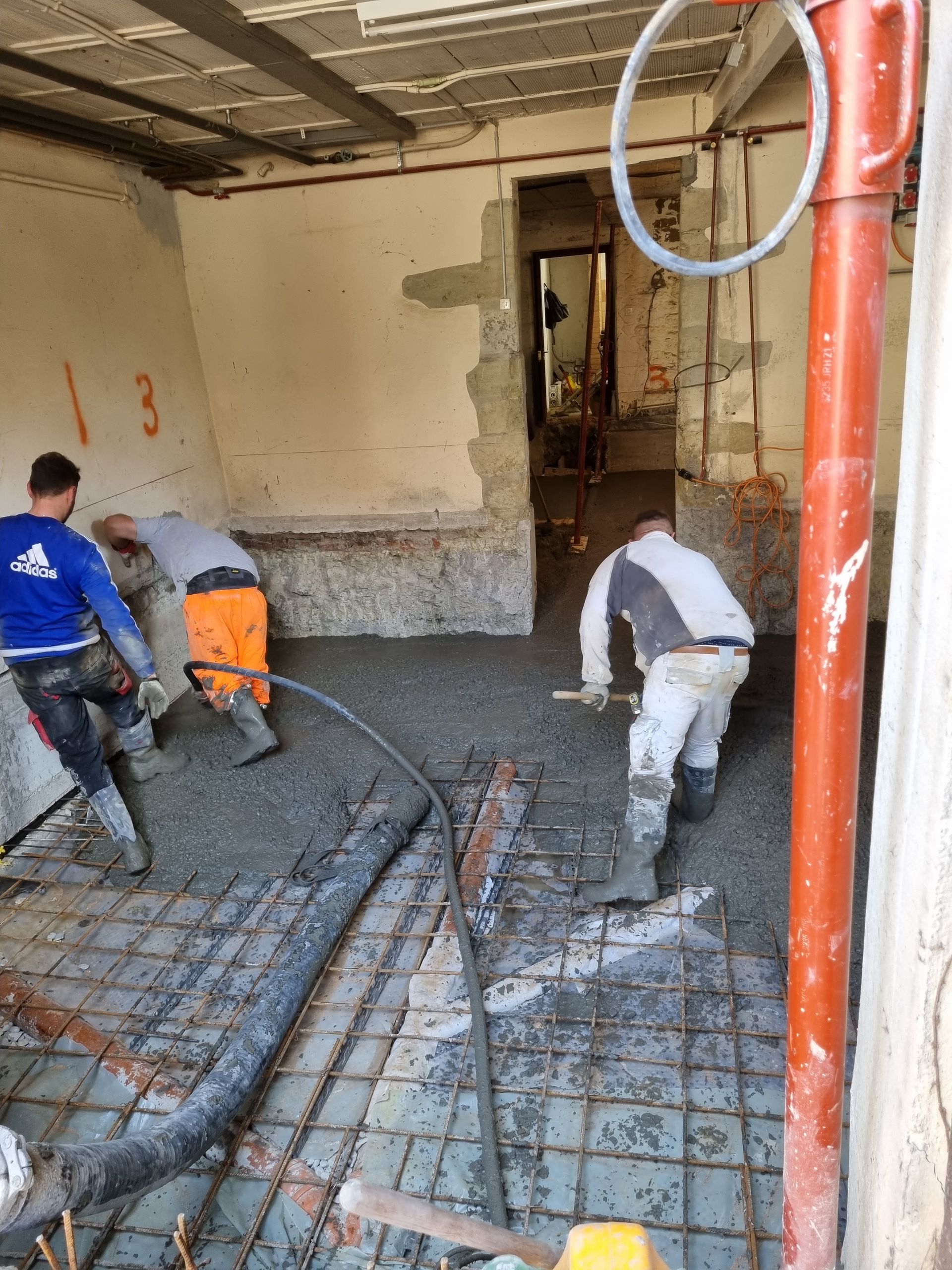 Trois ouvriers du bâtiment coulent et lissent du béton frais dans une pièce en rénovation, sur une grille métallique.