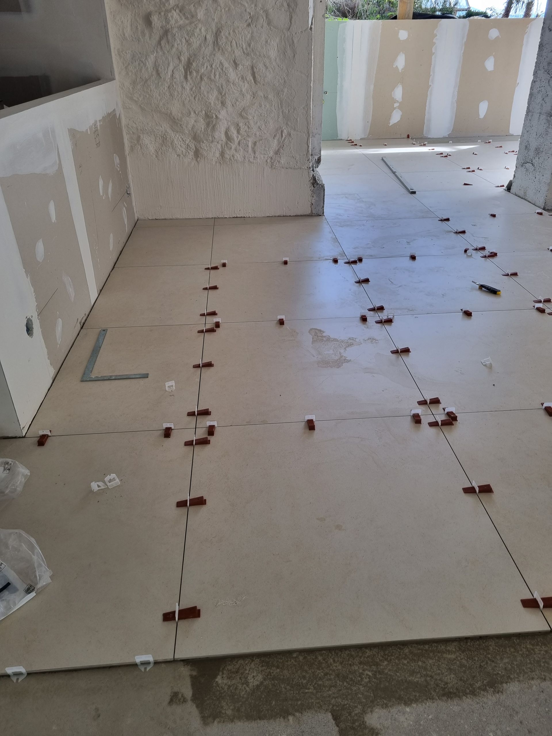 De grands carreaux de sol beiges ont été posés dans une pièce non finie, avec des croisillons en plastique marron et des murs recouverts de plaques de plâtre.