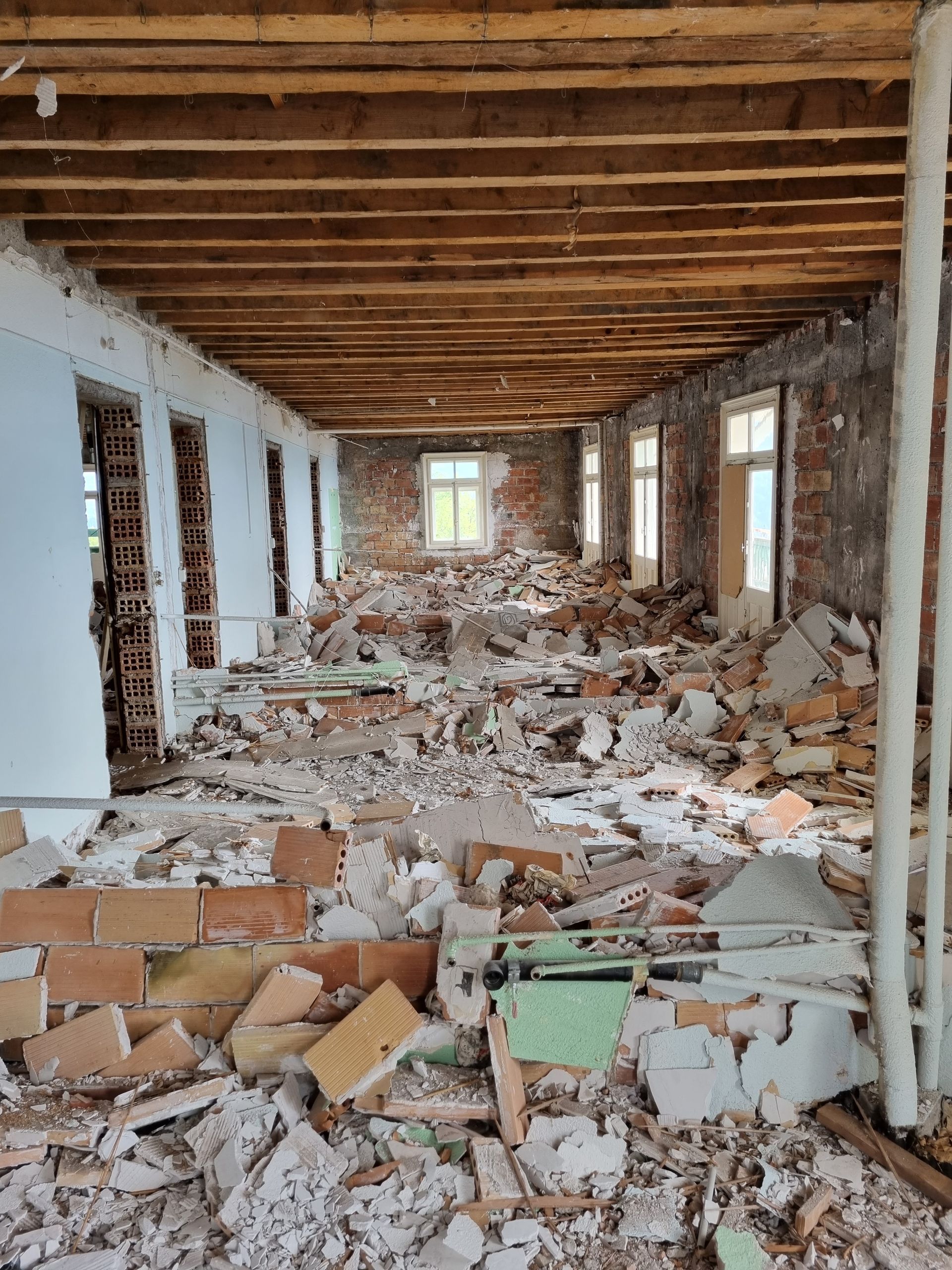 Une longue pièce intérieure rectangulaire en cours de démolition, remplie de débris de construction, de briques cassées et de solives apparentes.