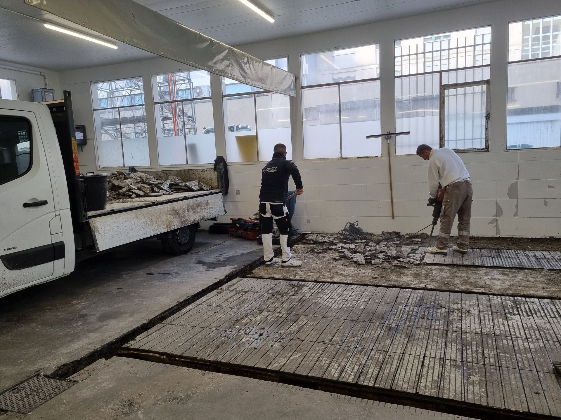 Deux ouvriers utilisent un marteau-piqueur pour démolir un carrelage à l'intérieur d'un bâtiment, à côté d'un camion de service blanc.