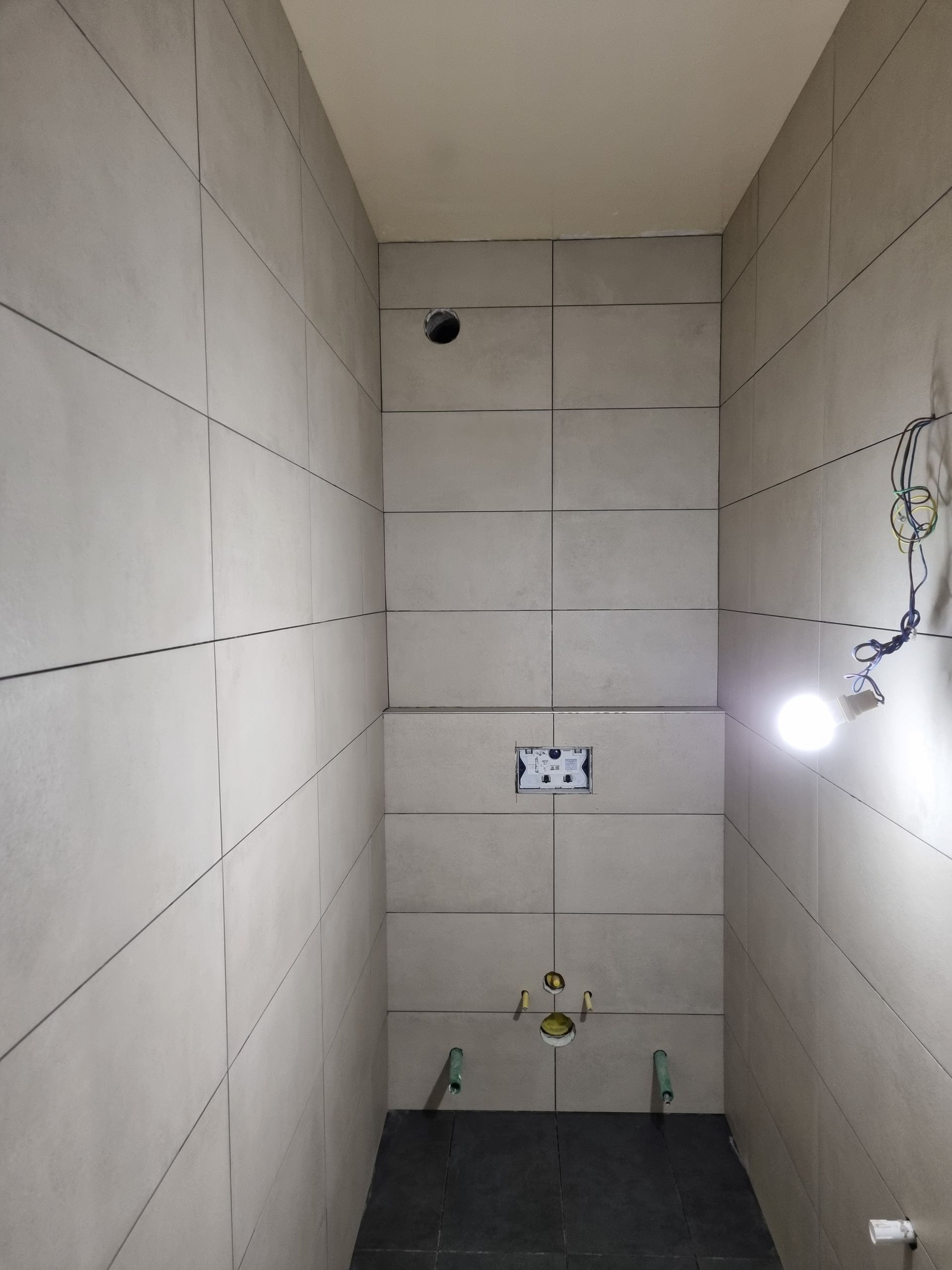 Une petite pièce étroite en construction, avec des murs carrelés de gris, un cadre pour l'installation de toilettes suspendues et un sol foncé.