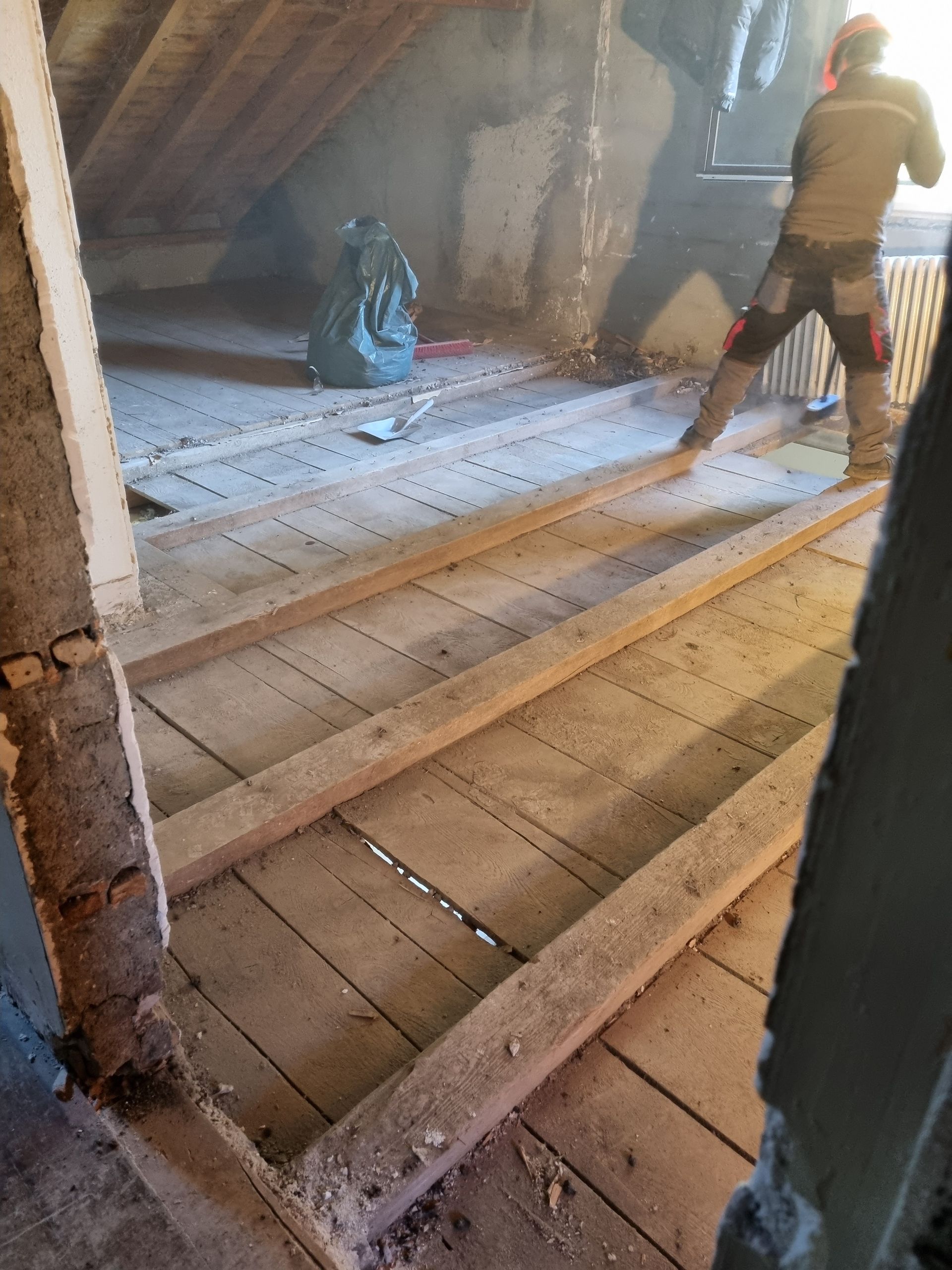 Un ouvrier en tenue de sécurité se tient debout sur des solives en bois lors de la rénovation d'un grenier, un sac à gravats vert se trouvant à proximité.