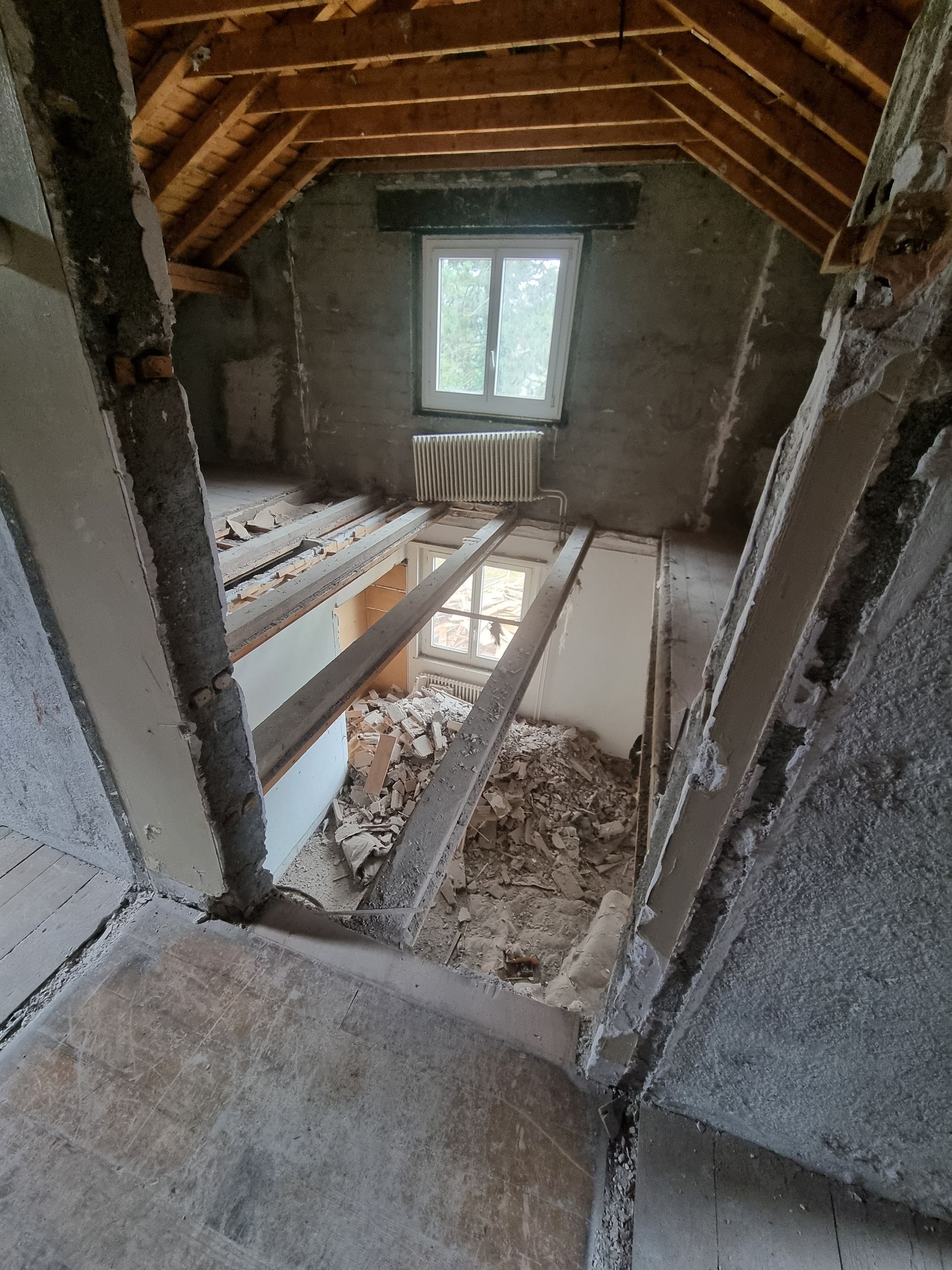 Une pièce mansardée inachevée avec des poutres en bois apparentes, une fenêtre et une grande partie du plancher ouverte laissant apparaître des débris en dessous.