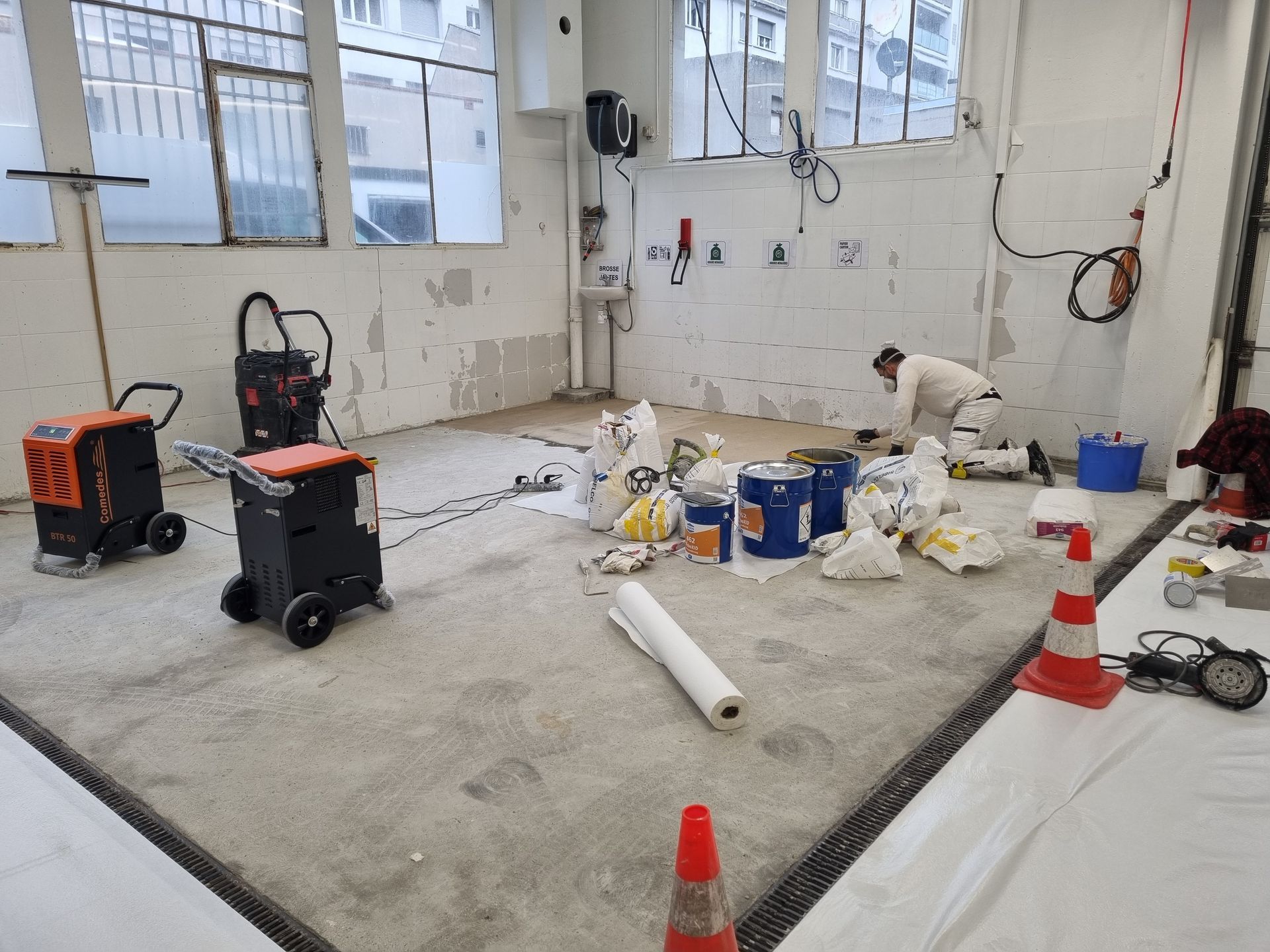 Un ouvrier en tenue de travail blanche est agenouillé sur un sol en béton dans une pièce où se trouvent du matériel de construction, des outils et des seaux de peinture.